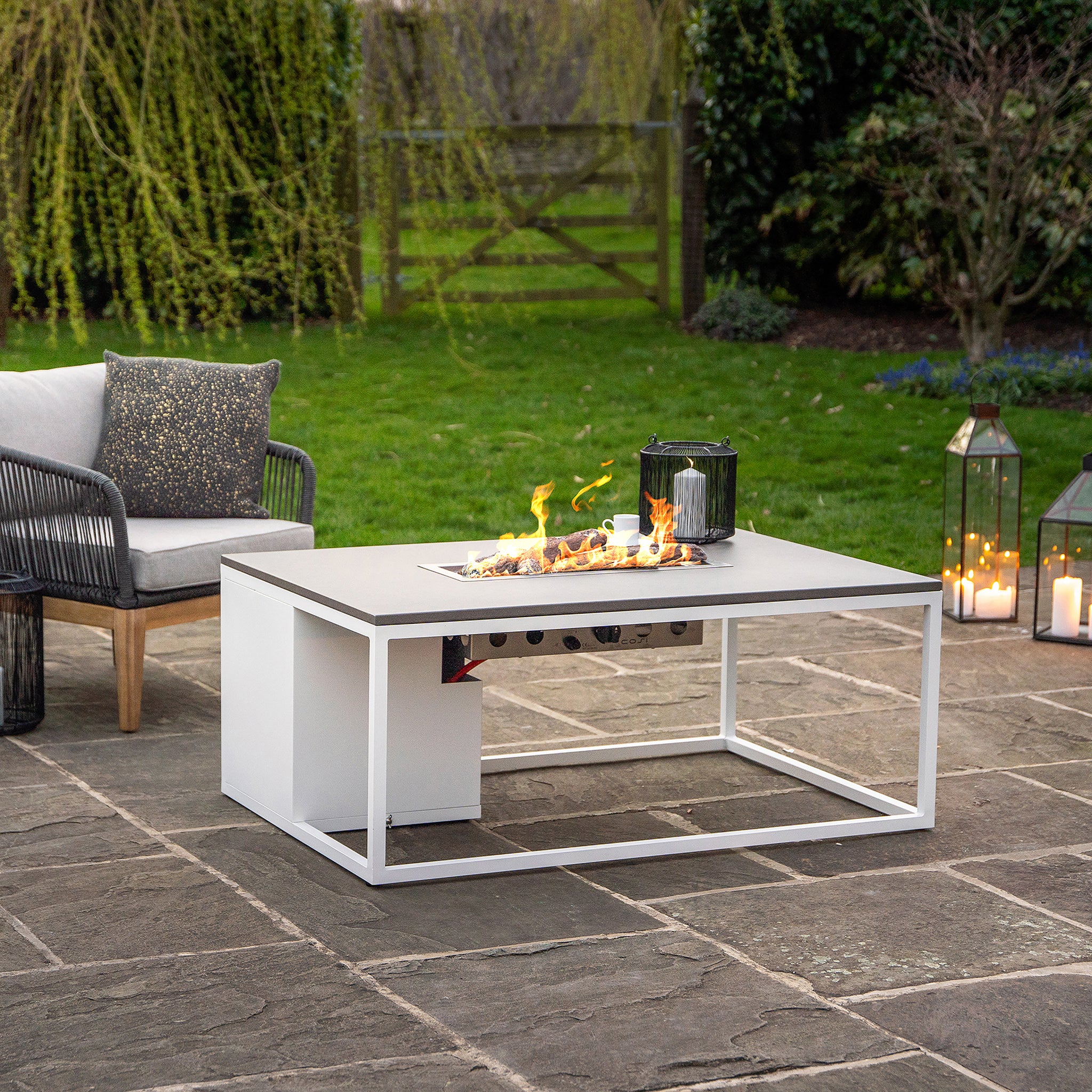 A modern outdoor lounging area featuring a Cosiloft 120 White and Grey Fire Pit Table, surrounded by chairs, lanterns, and candles on a stone patio in a garden.