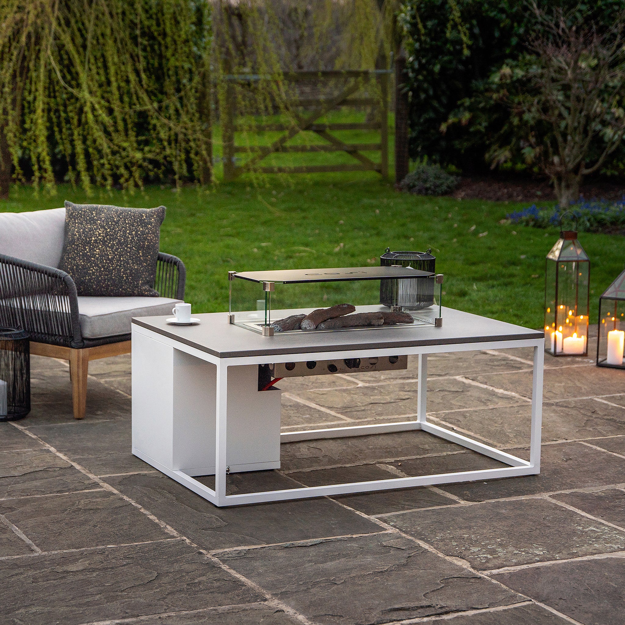 Modern outdoor patio featuring the Cosi - Cosiloft 120 White and Grey Fire Pit Table, a wicker sofa, and lanterns on a stone-tiled floor creates an ideal lounging space.