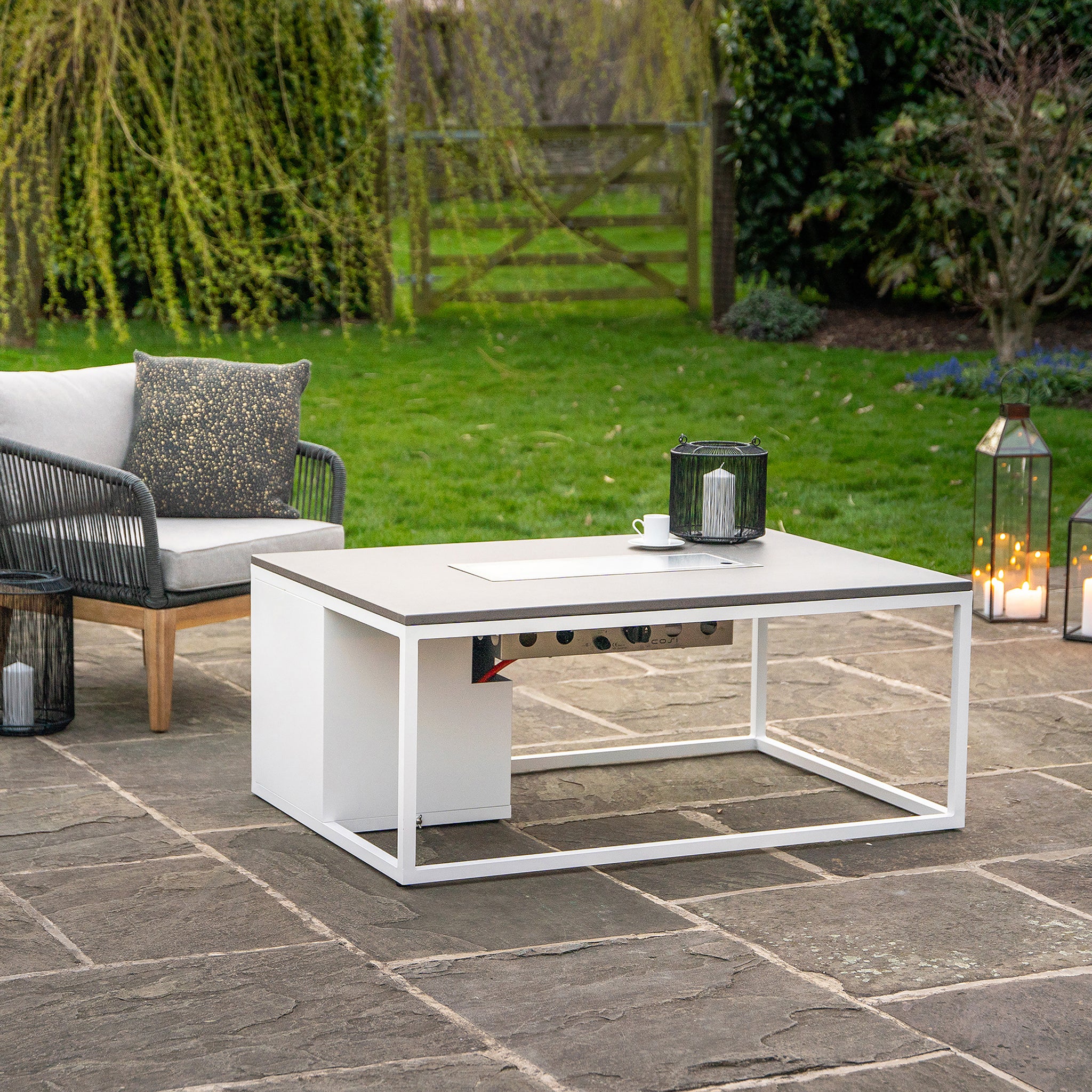 A modern outdoor patio features the Cosi Cosiloft 120 White and Grey Fire Pit Table, a sleek sofa, and greenery. Candles and a lantern on the concrete-style floor create a chic lounging vibe.