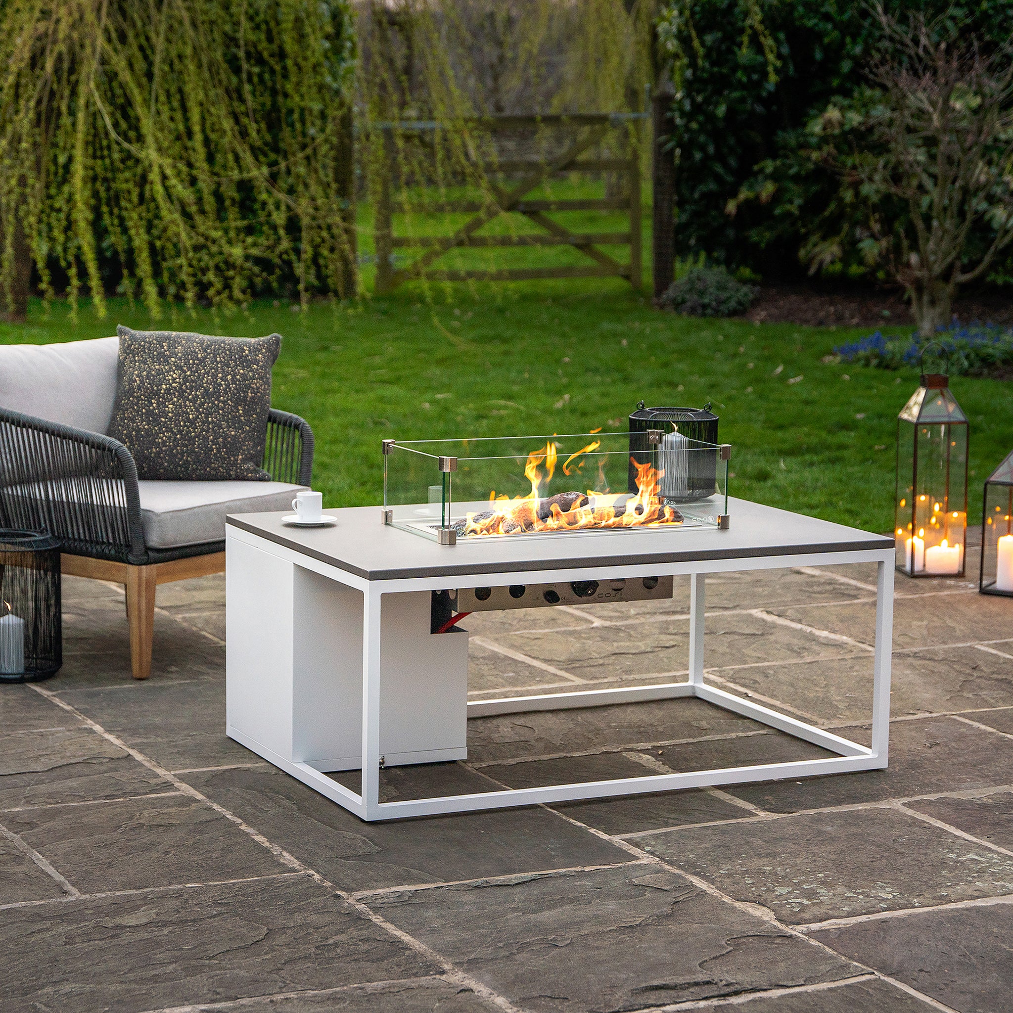 Modern patio with a Cosiloft 120 White and Grey Fire Pit Table, cushioned chairs, lanterns, and greenery in the background for a cozy outdoor lounging area.