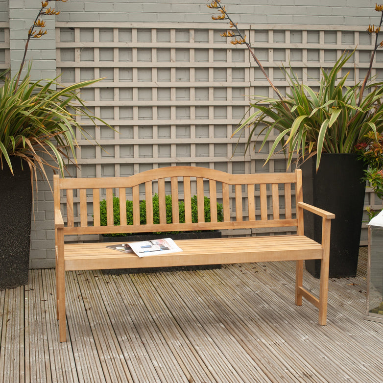 A Richmond Antique Wood Bench with Pop Up Table in Light Teak on a deck, flanked by two potted plants on each side, with an open book placed on the bench.