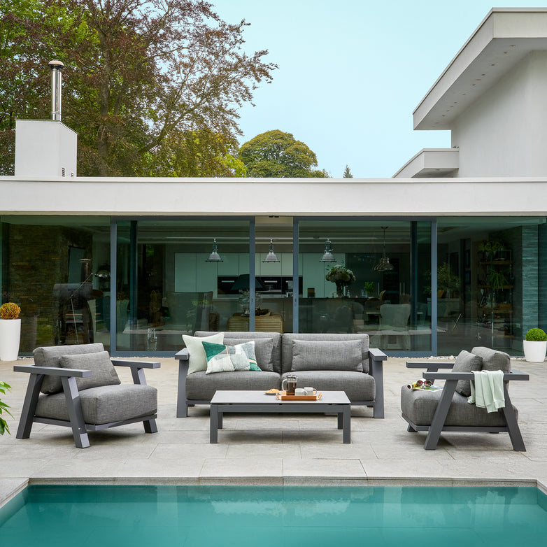 Modern outdoor patio with iconic outdoor furniture, featuring an anthracite 2 seat sofa set with weatherproof cushions and a Capitol HPL Coffee Table, beside a pool in front of a glass-walled house surrounded by trees.