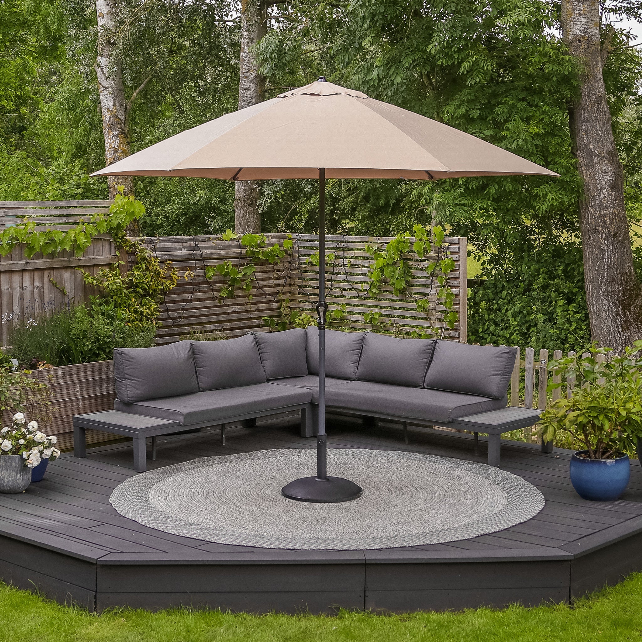 Outdoor patio with a gray sectional sofa, a round rug, and a khaki Triton 3m Round Aluminium Parasol offering UV protection and fade resistance, all surrounded by wooden fences and lush greenery.