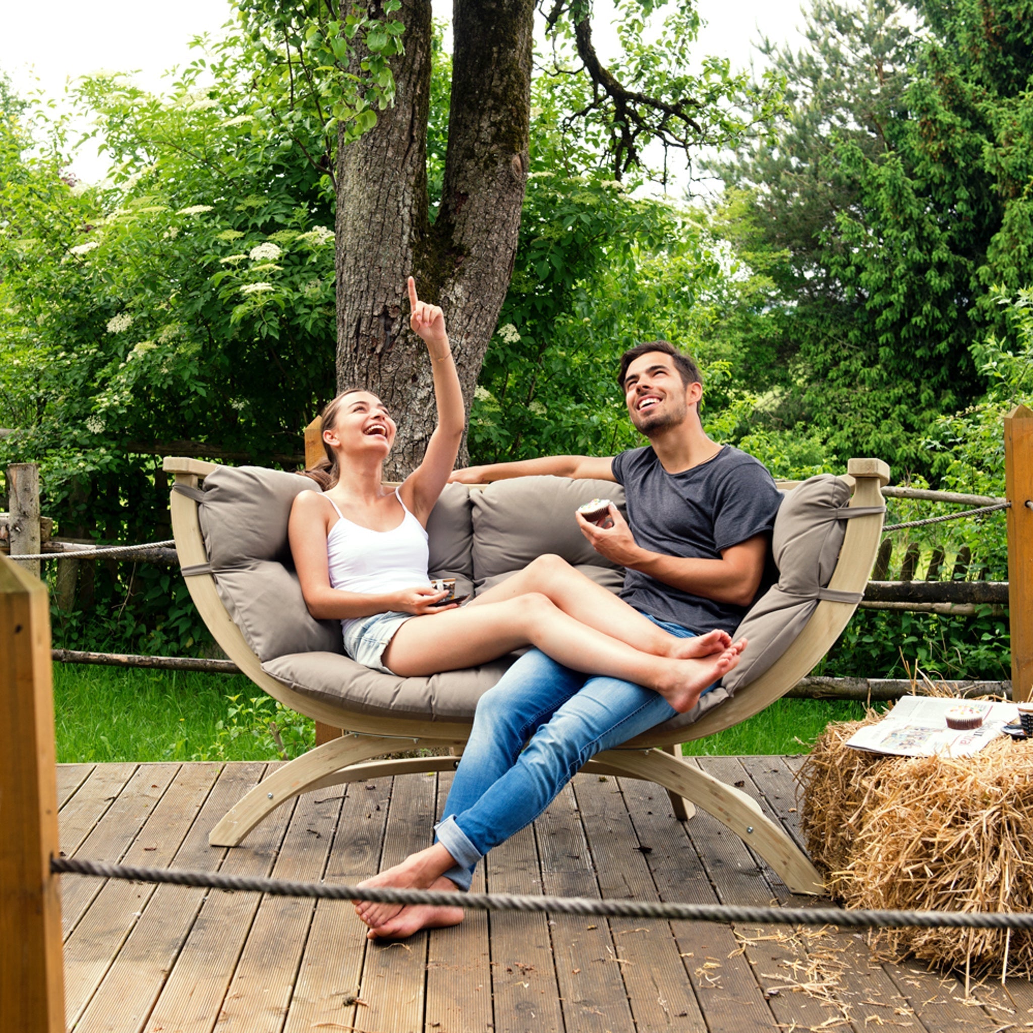 A couple sits laughing on a cozy, elegant Globo Siena Due Seat Weatherproof in Taupe Silver under a tree, surrounded by lush greenery and a bale of hay nearby. Crafted from sustainable wood, this versatile loveseat is perfect for outdoor-indoor use.