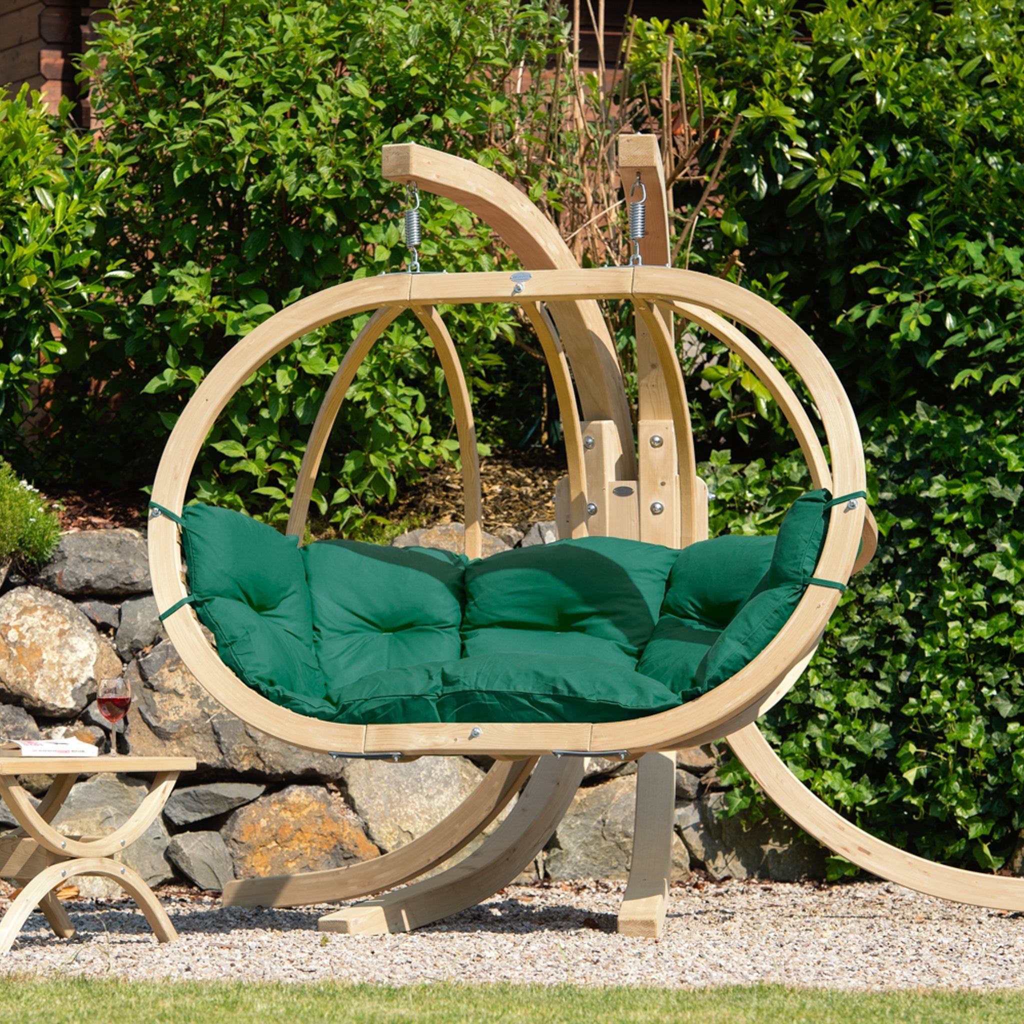 The Globo Royal Double Seater Hanging Chair Set, featuring weatherproof spruce wood and a Verde Green cushion, is perfectly set in a garden with lush greenery and a stone wall backdrop, secured with galvanized metal fixings.