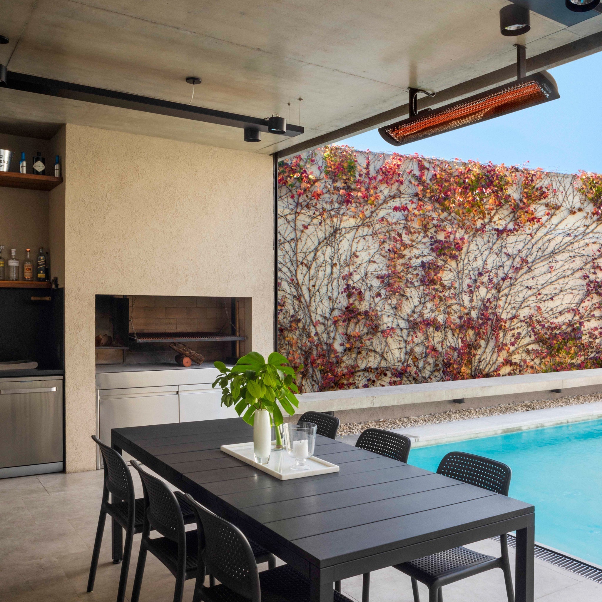 Outdoor dining area with a black table and chairs next to a barbecue, beside a swimming pool and a vine-covered wall, enhanced by the Bromic Tungsten Smart-Heat™ Electric 4000W in Black for comfortable evenings.