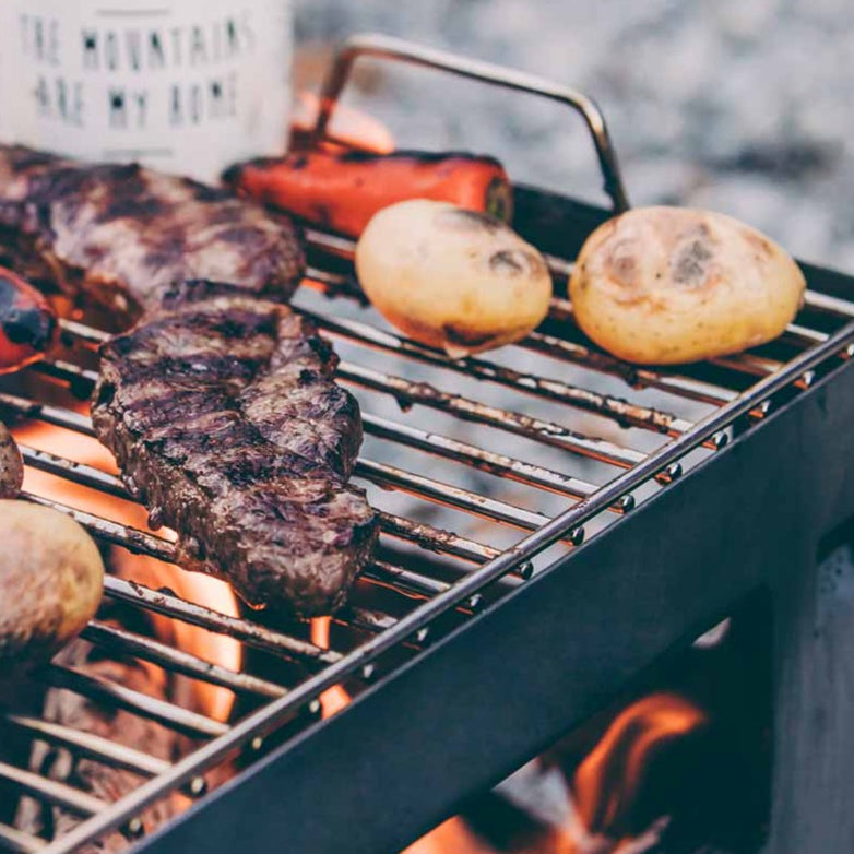 Grilled steaks, potatoes, and a red pepper on a barbecue grill with a "The mountains are my home" mug in the background. The Höfats BEER BOX Cooking Grid perfectly accommodates your feast.