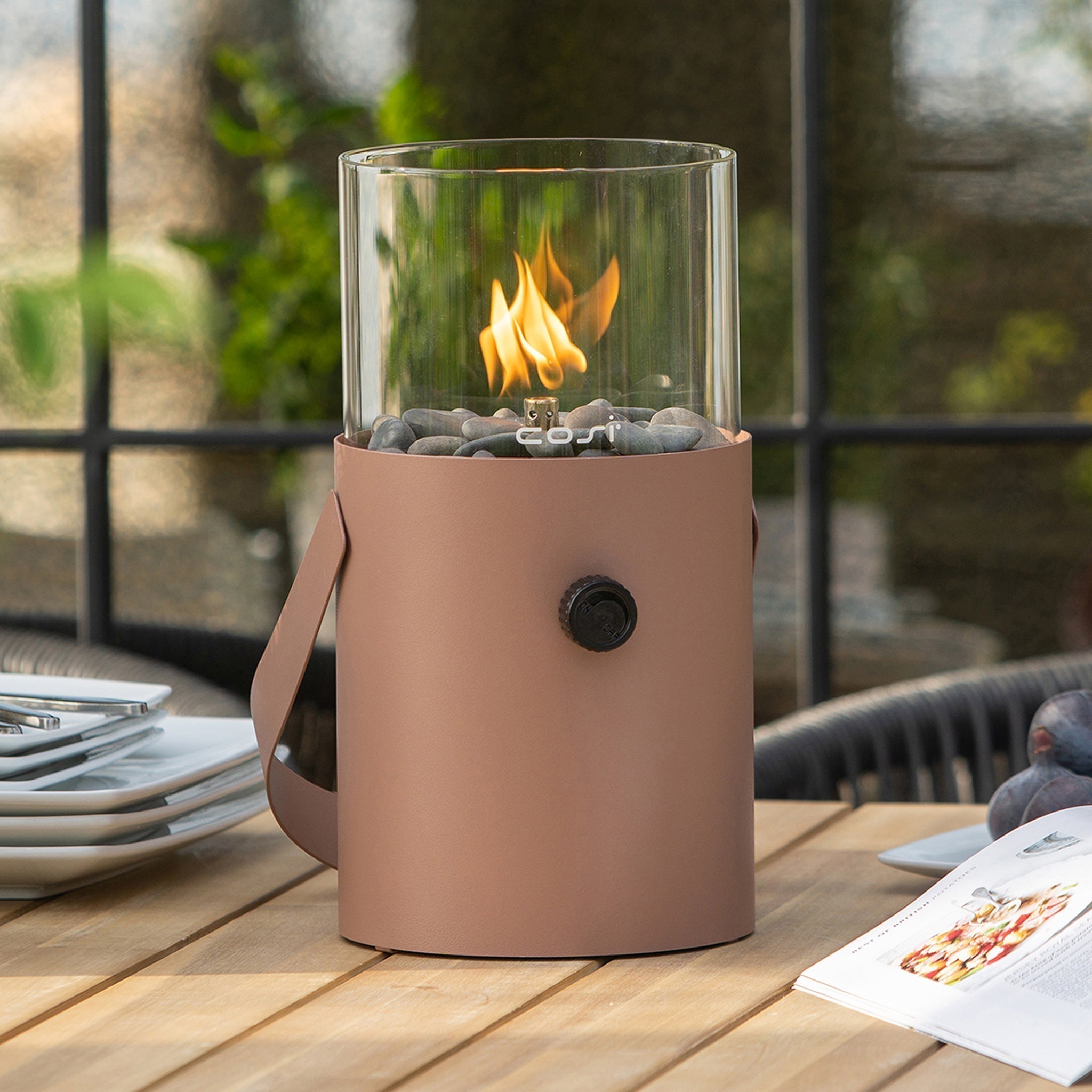 The Cosiscoop Fire Lantern in Terracotta, with its glass enclosure and terracotta base, placed on an outdoor wooden table near a window, adds charm to your garden lighting.