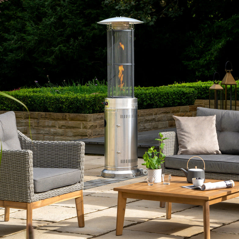 An outdoor patio with wicker furniture, a wooden table with a teapot, and the Pacific Lifestyle Cylinder Patio Heater in stainless steel elegantly displaying its flame.