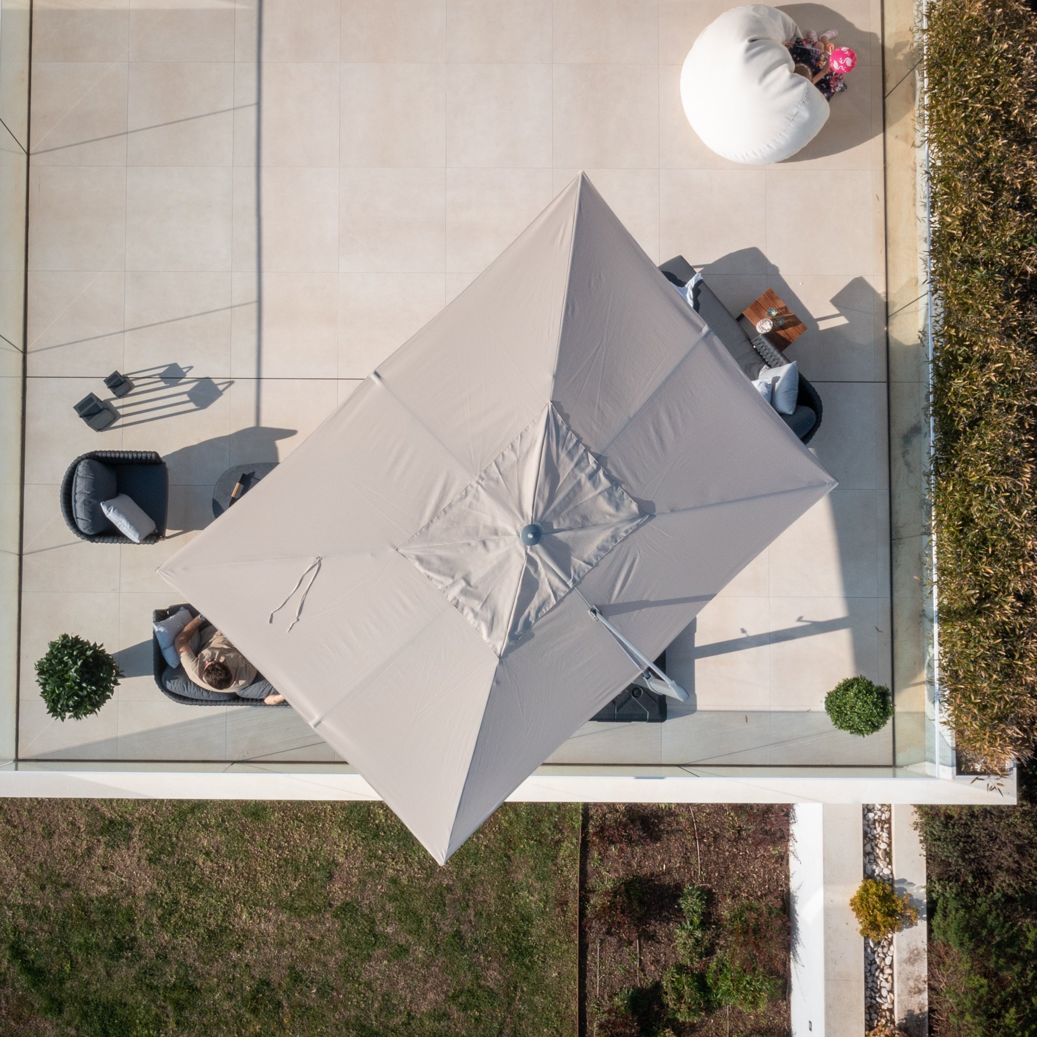 Top-down view of a patio featuring the Pallas 4m x 3m Rectangular Cantilever Parasol with LED Lighting in Beige, several chairs, and a bean bag near grass and shrubs, all beautifully illuminated by its subtle LED lights.