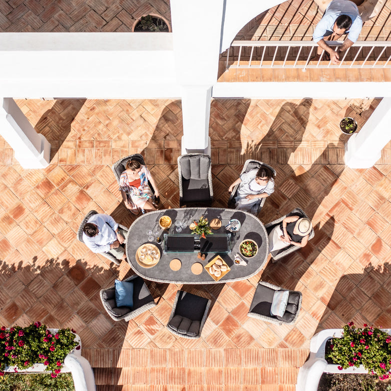 Aerial view of people sitting around a Santiago 8 Seat Rattan Oval Firepit Dining Set in Grey on a terracotta-tiled patio, with one person leaning on a balcony. This elegant dining set features aluminium powder-coated frames, adding both sophistication and durability to the outdoor setting.