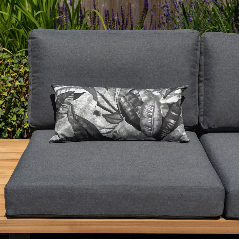 Gray outdoor couch adorned with Acrisol Amazonia Gris Bolster Scatter Cushions in black-and-white leaf patterns, placed on a wooden deck with plants in the background.