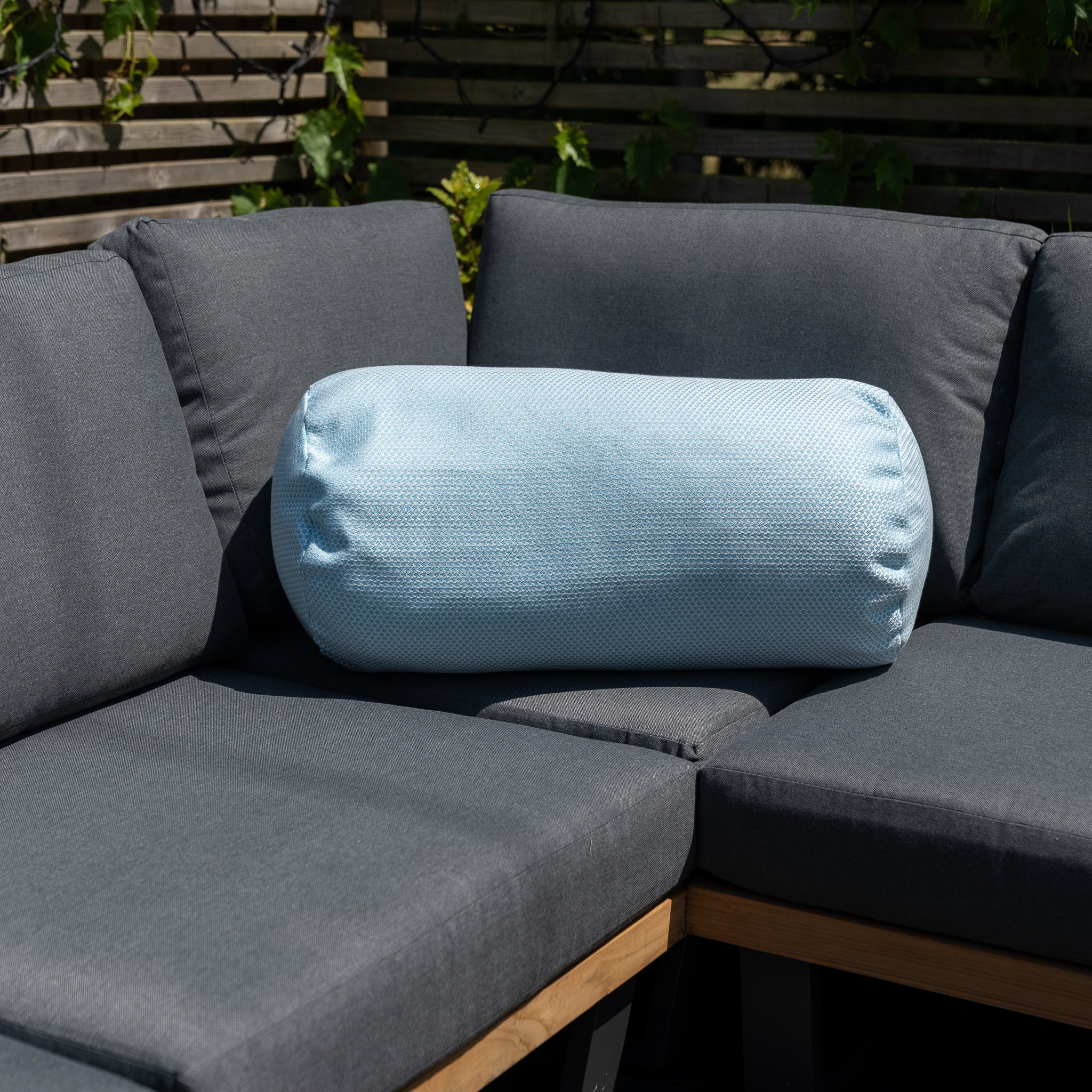 The Agora Scala Celeste Large Round Bolster Floor Cushion, measuring 72cm x 35cm and made from solution-dyed acrylic, rests on a dark gray outdoor sectional sofa with greenery in the background.