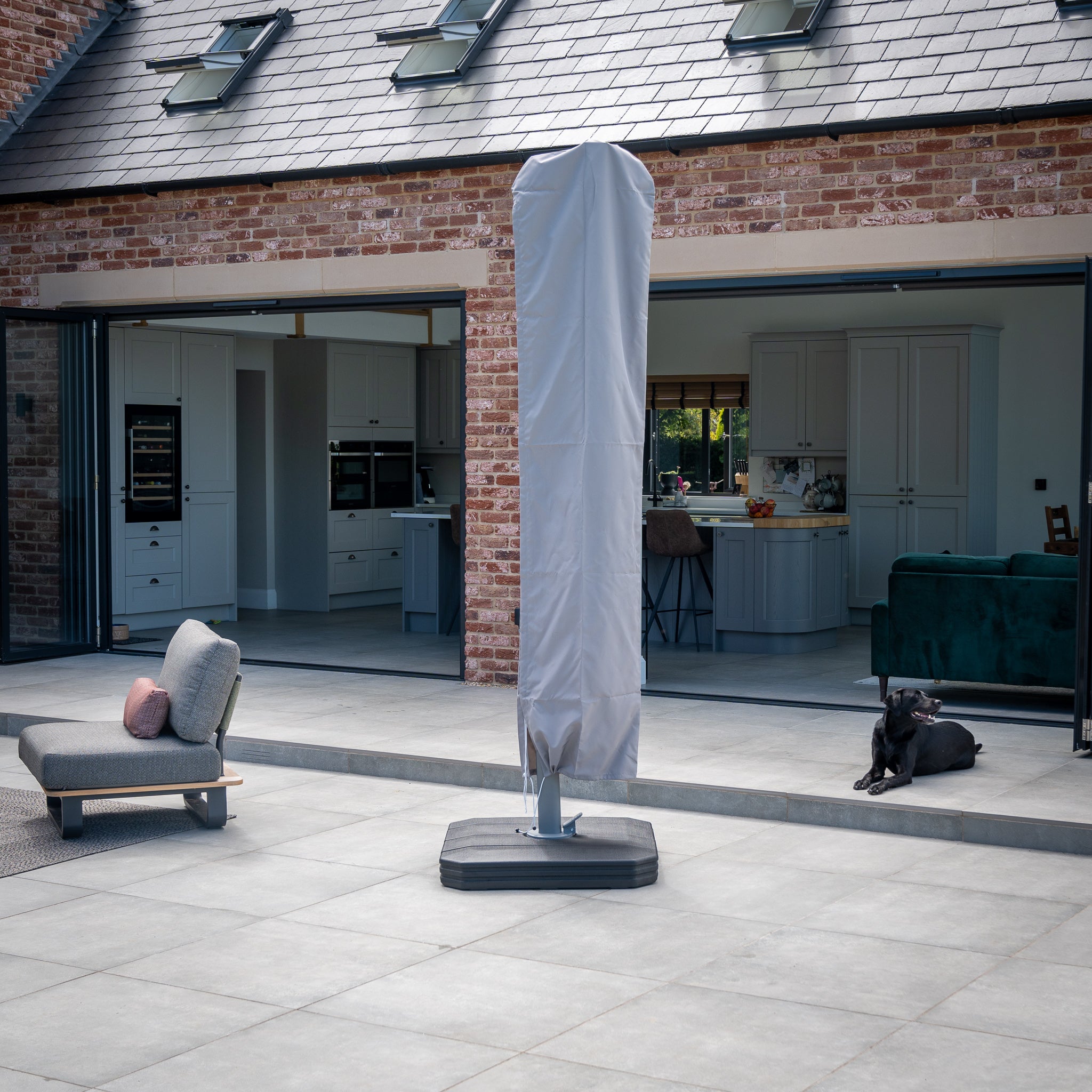 A Pallas 4m x 3m Rectangular Cantilever Parasol in Beige with LED lighting shades a black dog lying on a patio, with an open view of a modern kitchen in the background.