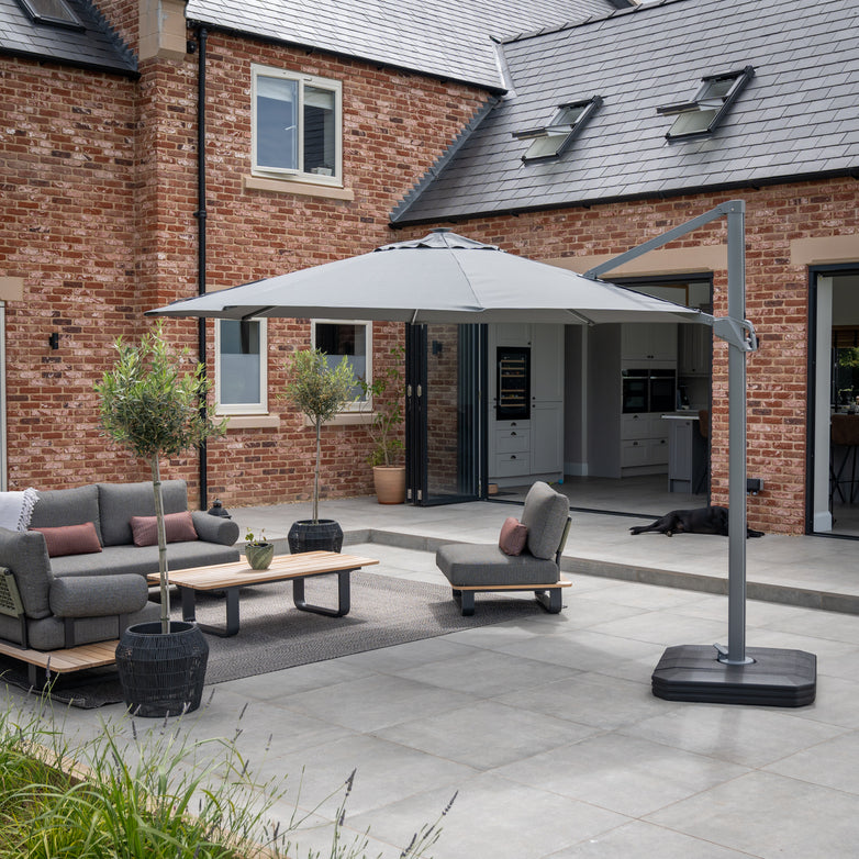 Patio with grey furniture and large umbrella, brick house with open glass doors in background, potted plants around. A sleek Ares 3.5m Round Cantilever Parasol with Solar-powered LED Lights in Charcoal completes the serene outdoor setting.