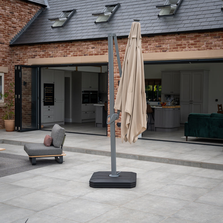 A folded Ares 3m Square Cantilever Parasol with a UV50+ acrylic canopy in beige stands on a tiled patio in a modern courtyard, illuminated by solar-powered LED lights, with open doors revealing an indoor space.