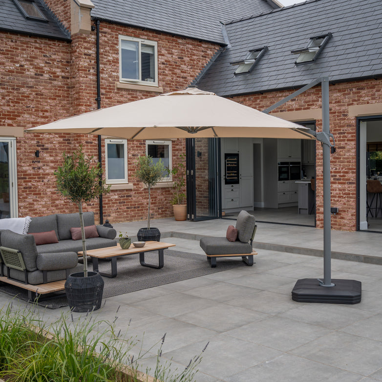 Modern patio with outdoor furniture under an Ares 3m Square Cantilever Parasol with Solar-powered LED Lights in Beige, featuring a UV50+ acrylic canopy. This elegant large umbrella stands tall, illuminating the space with LED lights powered by a Solar Charging Battery Pack, set against the backdrop of a charming brick house.