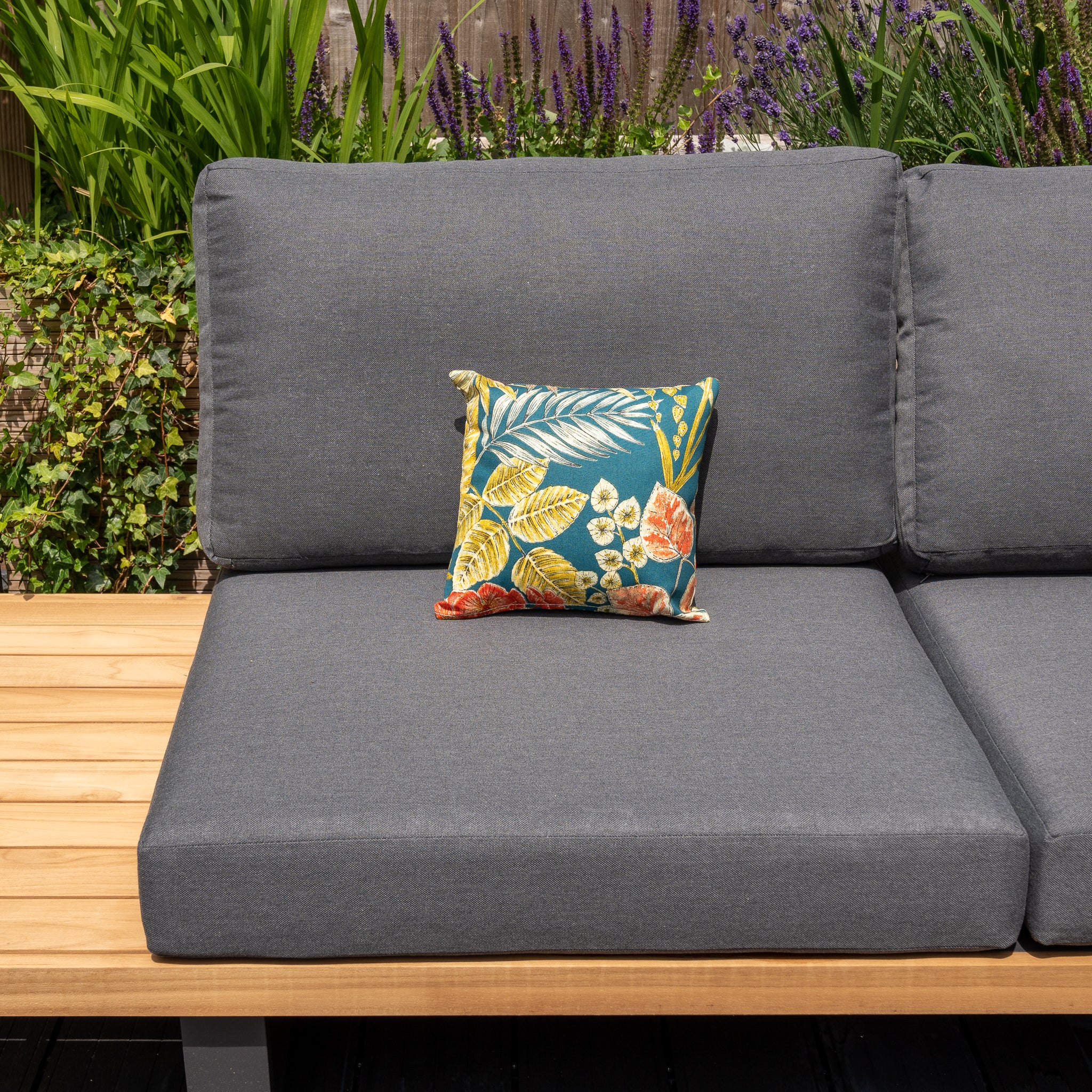 Gray outdoor couch with a wooden armrest and a colorful floral pillow, complemented by the Acrisol Bouquet Petrol Small Scatter Cushions (25cm x 25cm), set in front of lush green plants and purple flowers.