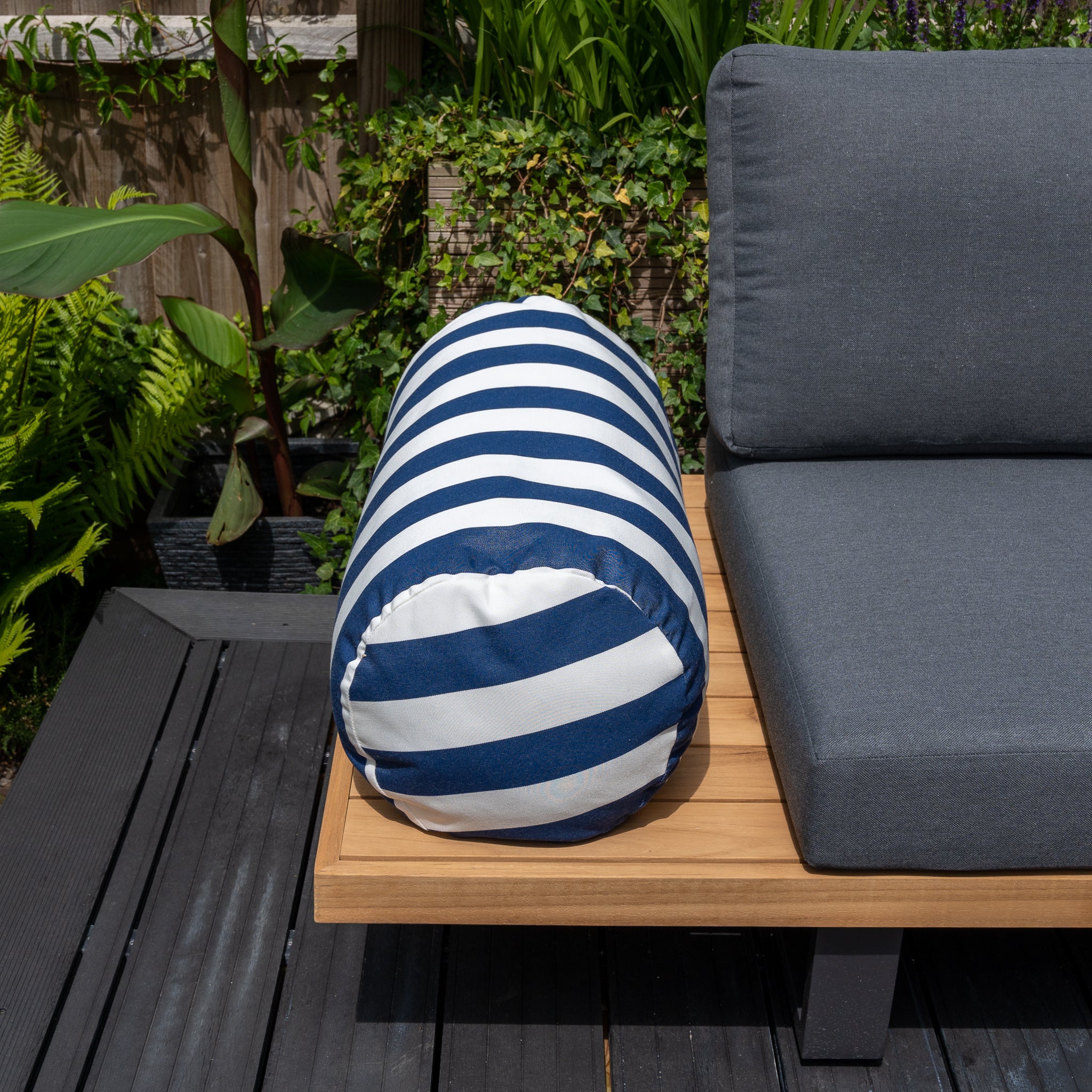 An Agora Lines Marino Large Round Bolster Floor Cushion (72cm x 35cm) sits on a wooden patio bench with grey cushions, surrounded by green plants and foliage, showcasing the durability of solution-dyed acrylic.