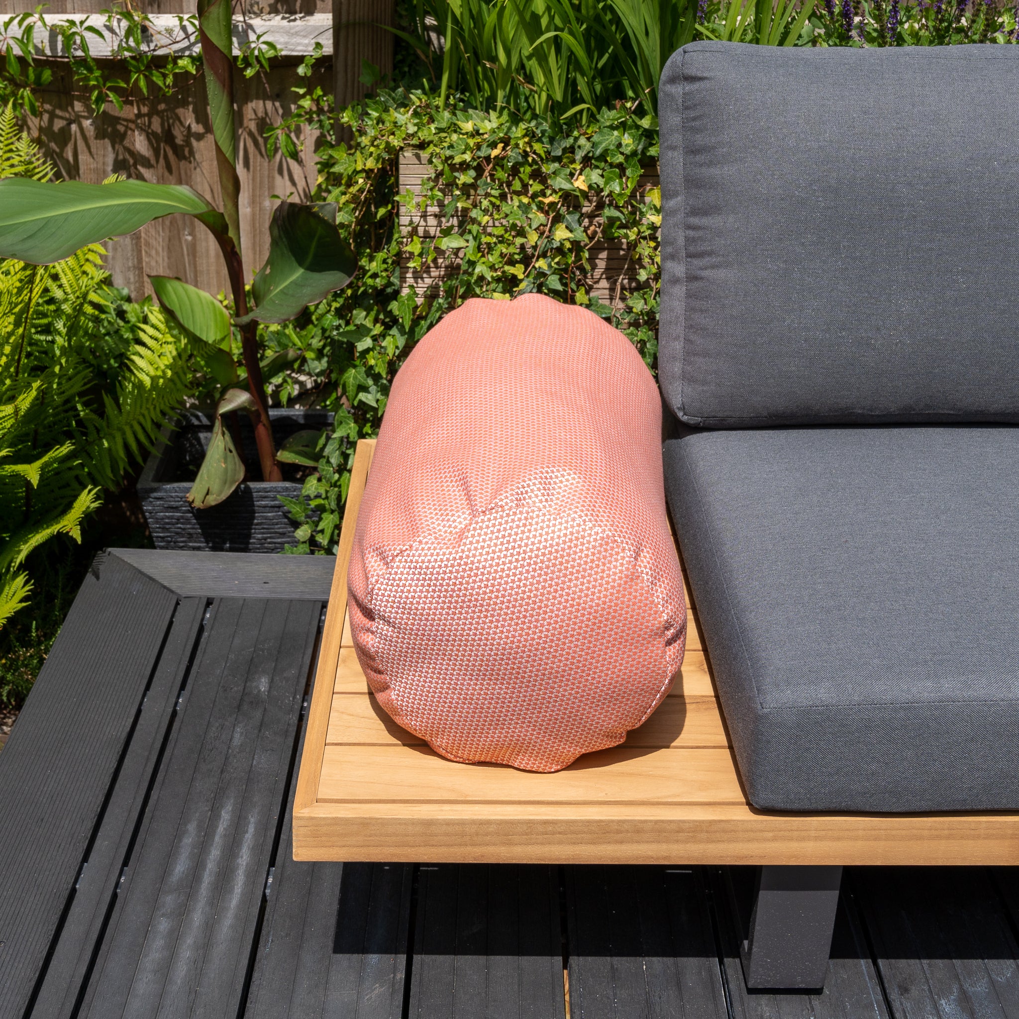 Outdoor seating featuring a gray cushion made from solution-dyed acrylic and the Agora Scala Coral Large Round Bolster Floor Cushion - 72cm x 35cm on a wooden bench, surrounded by lush green plants. Perfectly complemented by outdoor scatter cushions for added comfort.