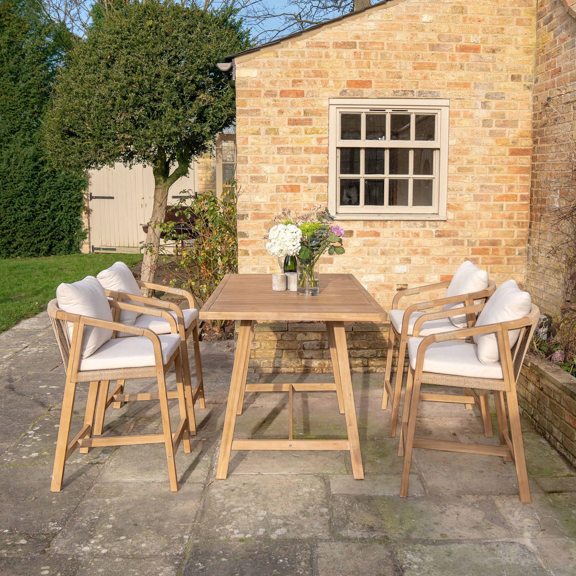 Patio with a Quay 4 Seat Rectangular Bar Set in Linen, crafted from sustainably sourced acacia wood, four cushioned chairs, flowers in vases, and a brick house in the background.