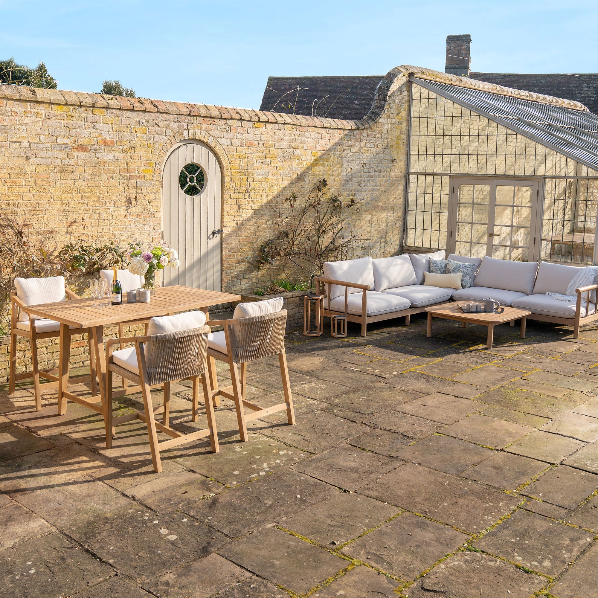 A spacious patio featuring the Lavenham Corner Sofa Set in Linen with beige cushioned wooden outdoor furniture crafted from sustainable eucalyptus wood, a small potted plant, and a yellow brick wall backdrop.