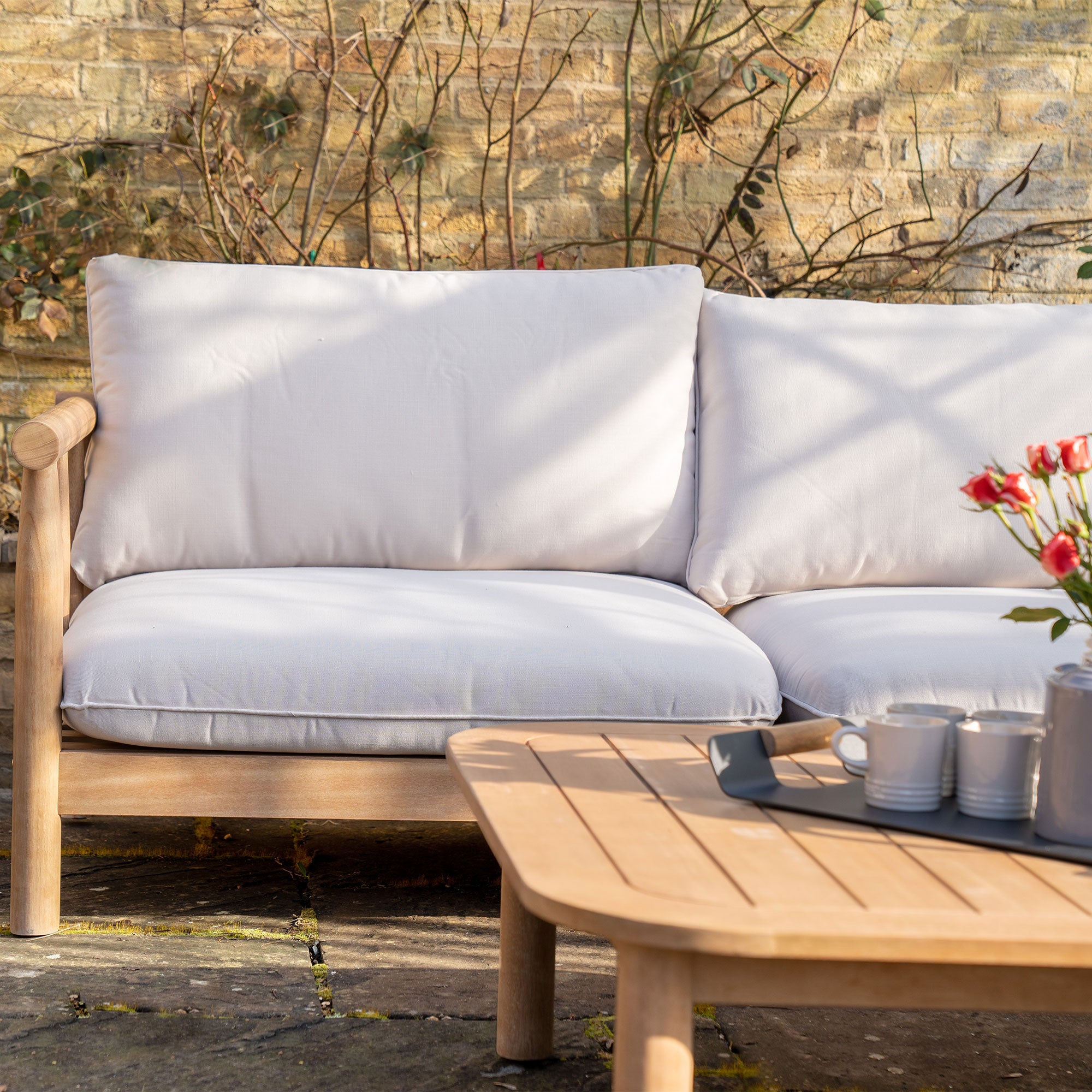 A cozy outdoor patio setup featuring the Lavenham Corner Sofa Set in Linen, complete with a cushioned wooden sofa, a coffee table with mugs, and flowers against a brick wall.