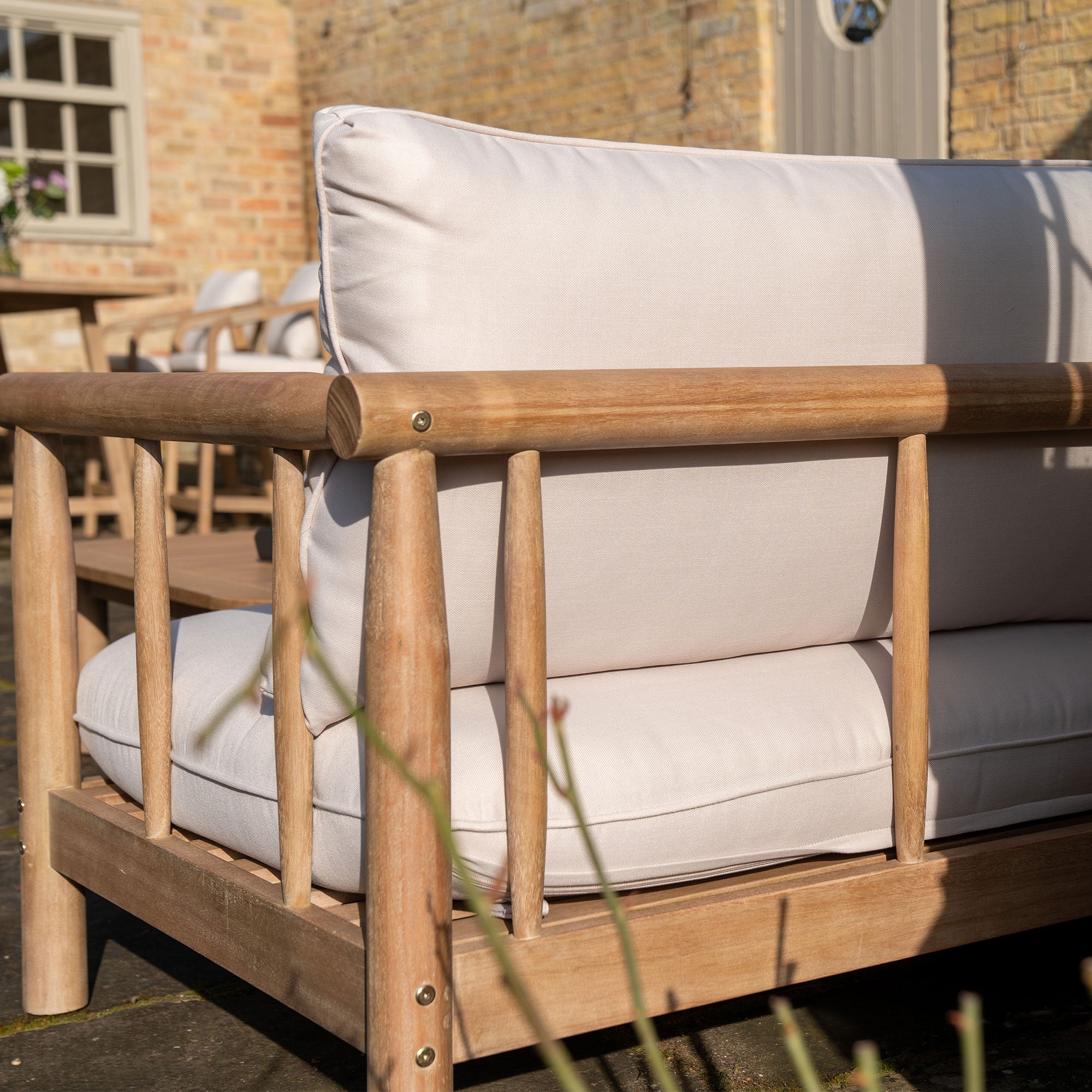 The Lavenham Corner Sofa Set in Linen, made from sustainable eucalyptus wood and featuring light beige cushions, is elegantly placed on a sunlit patio with a brick wall backdrop and a table with chairs in the distance.