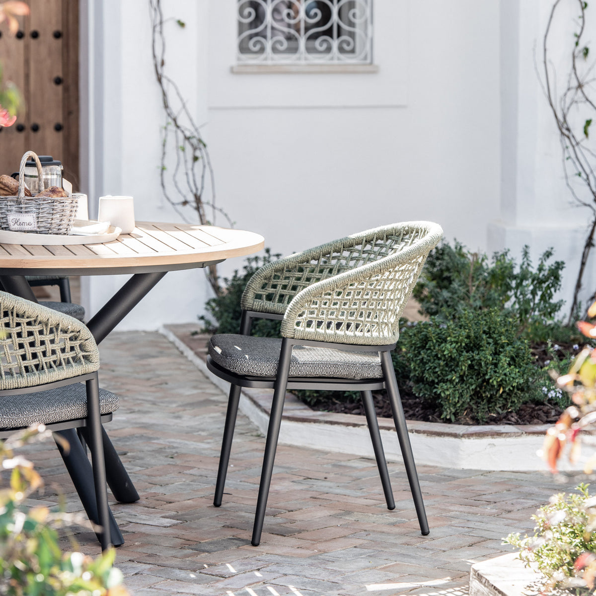 Kalama 4 Seat Round Dining Set with Teak Table in Olive Green, featuring two woven chairs with sun-proof cushions, situated on a brick patio amid lush greenery and framed by pristine white walls.