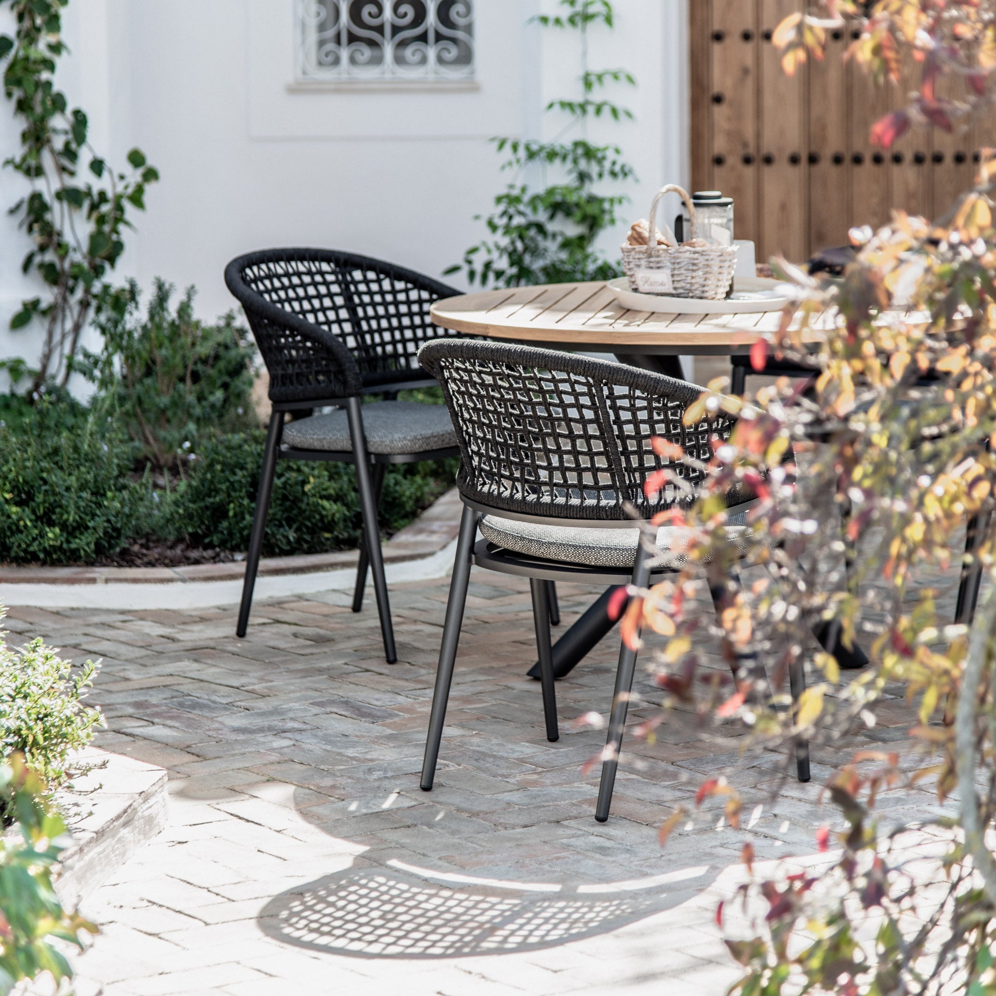 Outdoor patio featuring the Kalama 4 Seat Round Dining Set with a Teak Table in Charcoal, surrounded by greenery against a white wall.