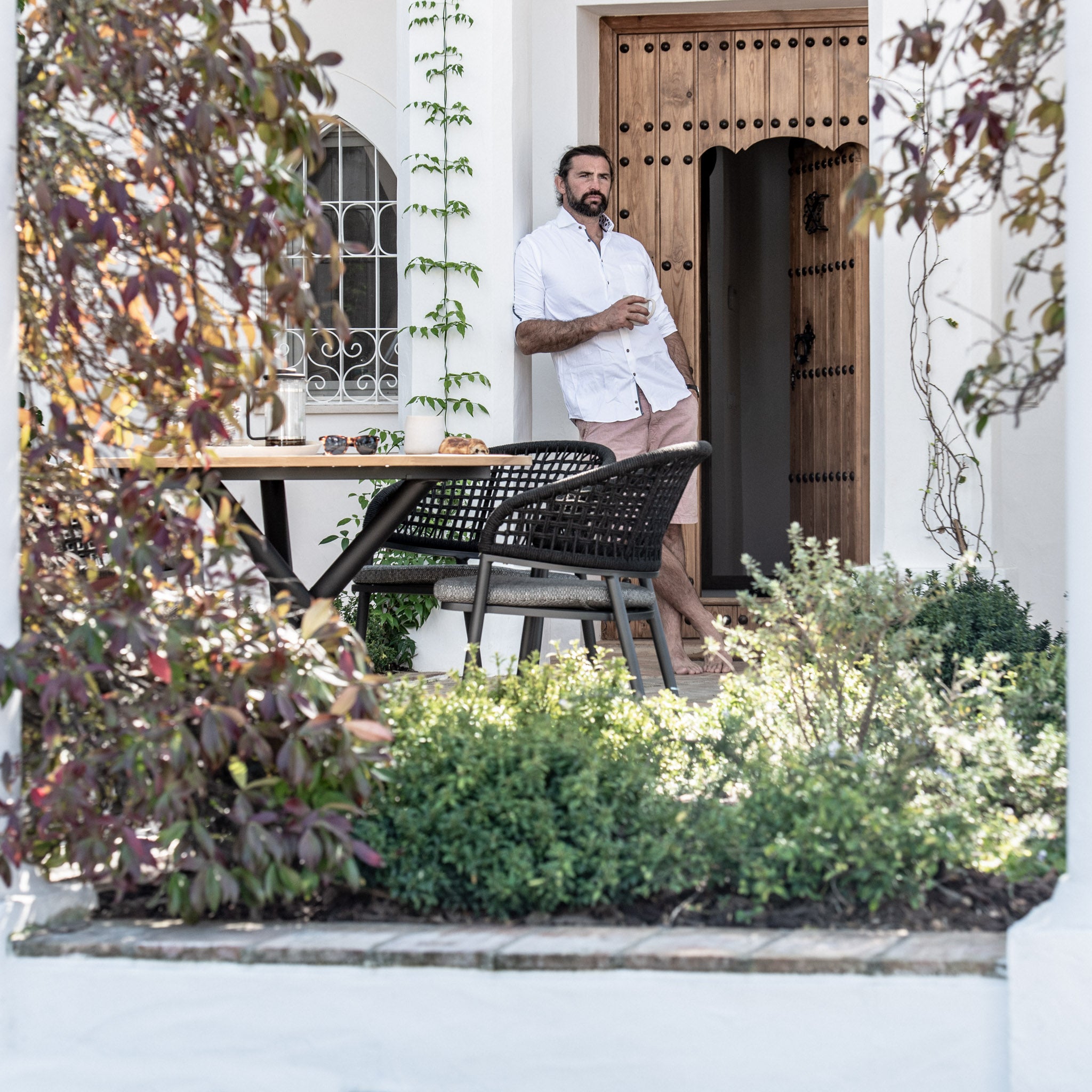 A man stands in a well-kept garden, holding a drink beside the Kalama 4 Seat Round Dining Set featuring a teak table in charcoal and woven chairs, with a wooden door in the background.