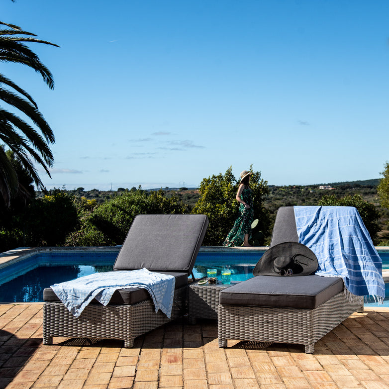 Two Santiago Rattan Sunloungers from the Santiago Rattan Sunlounger Set with Side Table in Grey are by a pool, one draped with a blue towel, under a clear blue sky. The high-quality reflex foam provides exceptional comfort while the adjustable backrests ensure personalized relaxation.