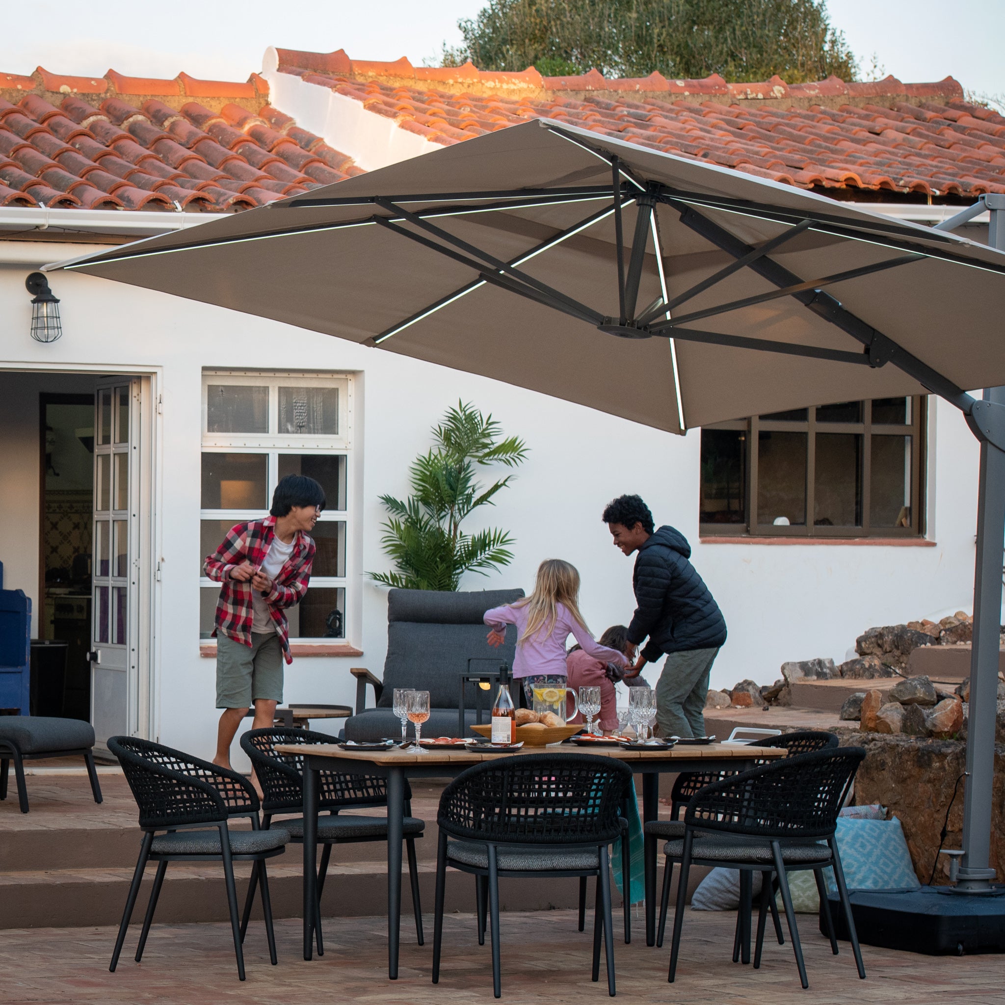 People enjoying an outdoor gathering under the Pallas 4m x 3m Rectangular Cantilever Parasol with LED Lighting in Beige near a house with a red-tiled roof.