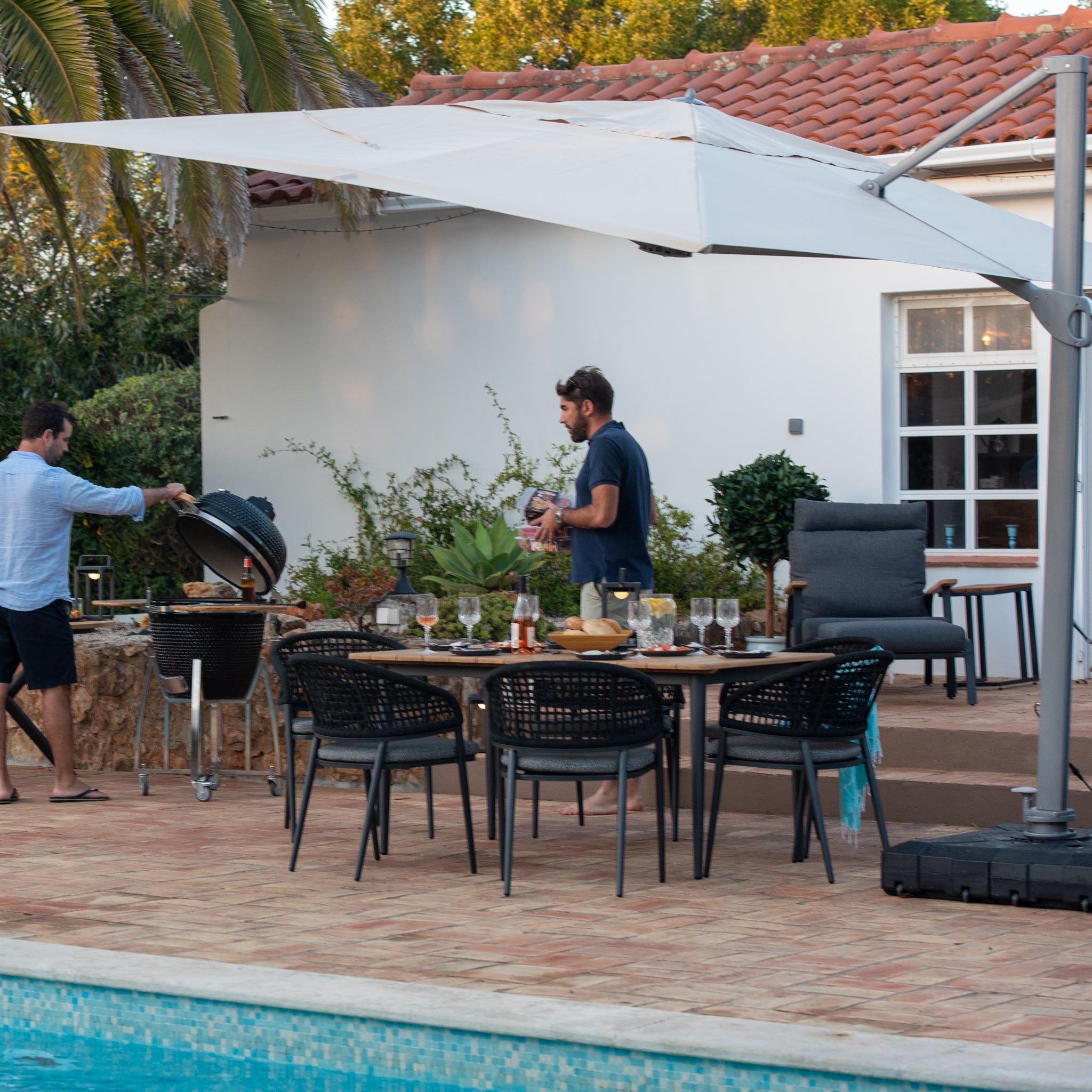 Two men preparing a meal by a poolside table under a Pallas 4m x 3m Rectangular Cantilever Parasol with LED Lighting in Beige, with a house and outdoor furniture in the background.