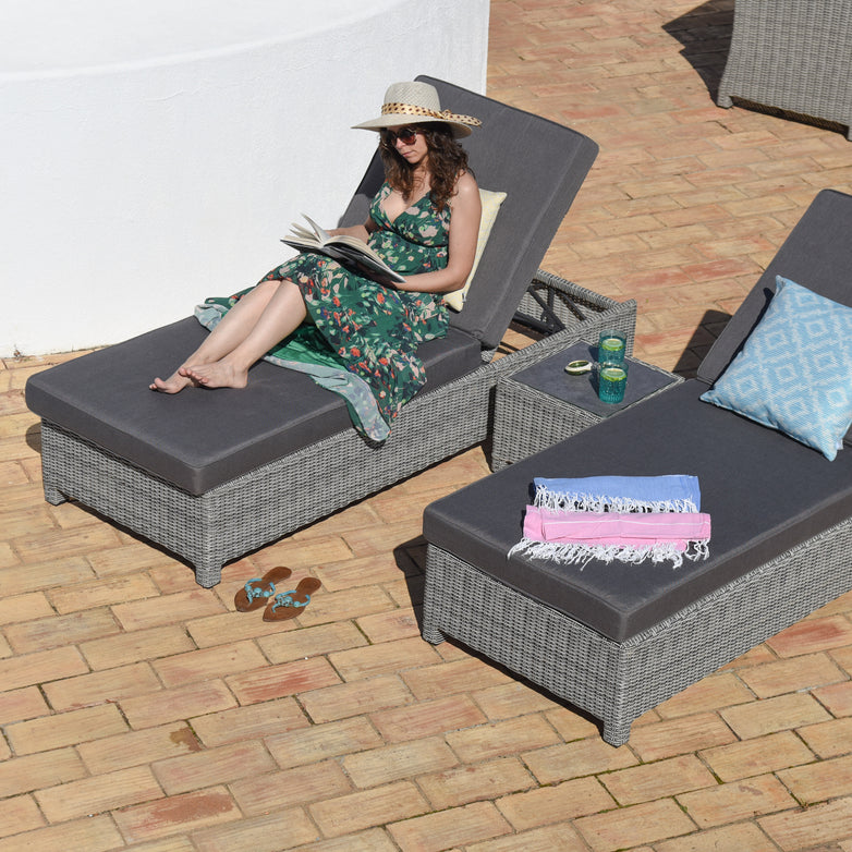 A woman relaxes on a Santiago Rattan Sunlounger Set with Side Table in Grey, reading in the sun while wearing a floral dress and hat. The outdoor setting is complete with high-quality reflex foam cushions and adjustable backrests, surrounded by complementary furniture and accessories.