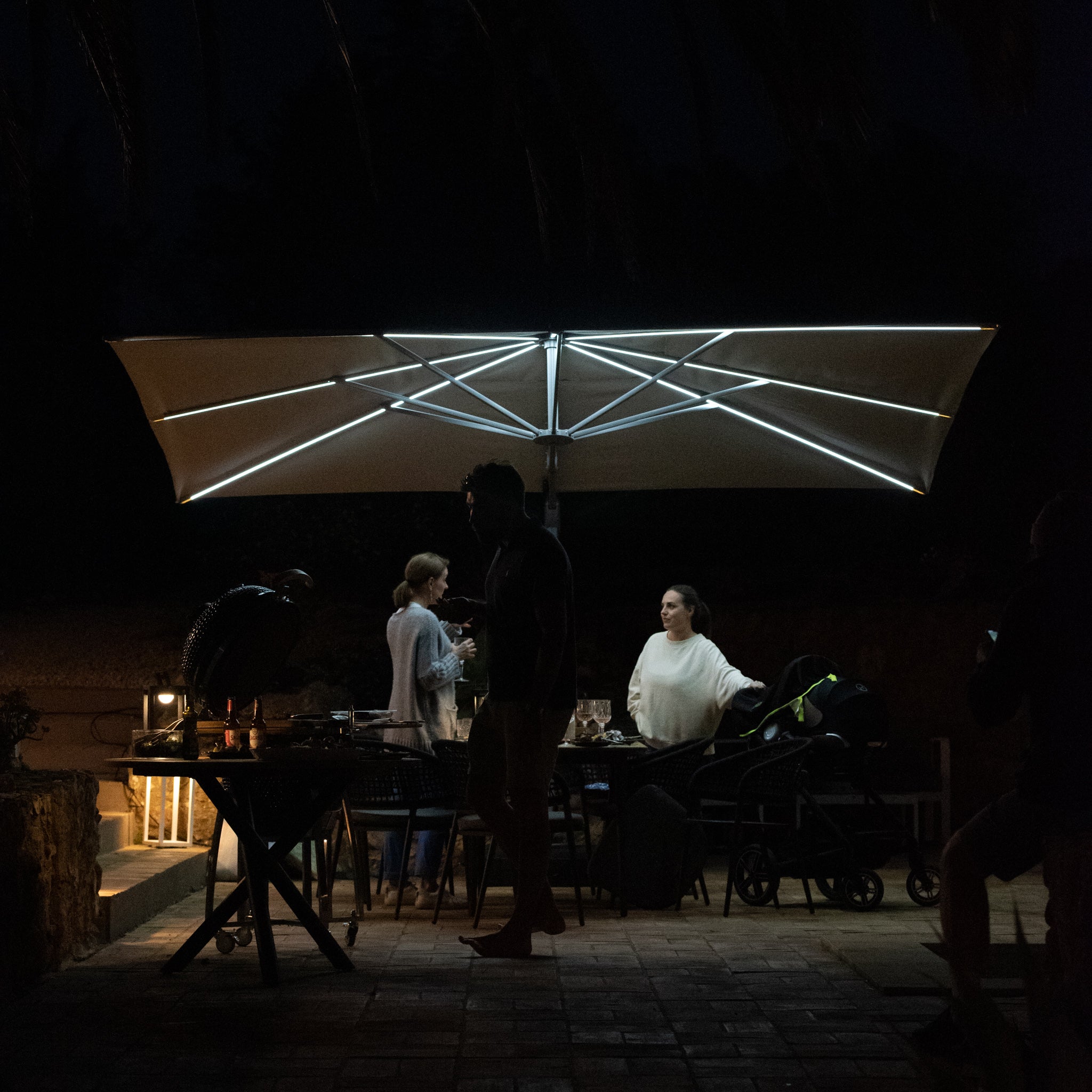 A group of people gathered under the Pallas 4m x 3m Rectangular Cantilever Parasol with LED Lighting in Beige at night, engaging in conversations and activities, illuminated by soft LED lights.