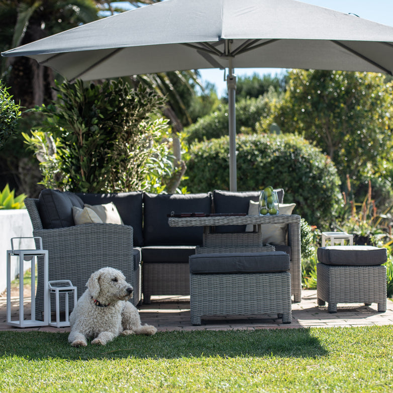 Outdoor patio set with umbrella, featuring the Santiago Square Rattan Corner Dining Set with Rising Table in Grey and gray-cushioned wicker furniture. The aluminum powder-coated frames ensure durability, and a white dog lies peacefully on the grass nearby.