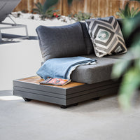 A modern outdoor sofa with gray cushions and a contemporary aluminium frame, accompanied by a wooden Panama Teak Side Table adorned with a blanket and magazine.