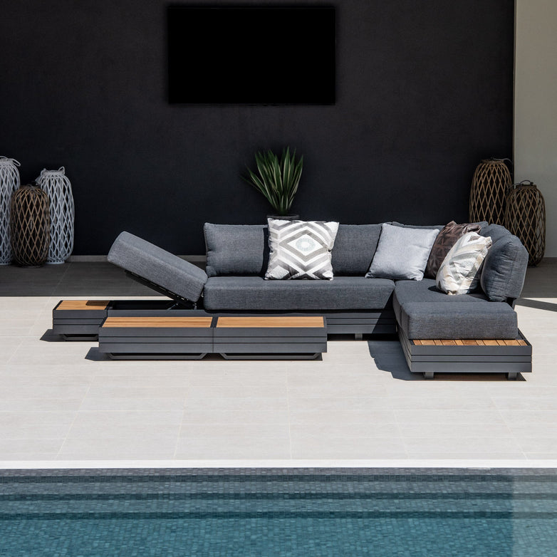 Modern outdoor patio featuring the Panama Luxury Outdoor Large Corner Group Set in Charcoal, complete with plush pillows and low coffee tables, beside a swimming pool, against a dark wall.