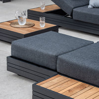 The Panama Corner Unit/Footstool in Charcoal offers modern outdoor seating with gray cushions, a wood and metal frame, and a wooden side table adorned with glassware. This modular system includes a versatile corner unit and an optional footstool for enhanced comfort.