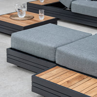 Gray cushioned outdoor seating with wooden side tables, featuring a glass pitcher and two glasses on a woven placemat, complemented by the versatile Panama Corner Unit/Footstool in Washed Grey.