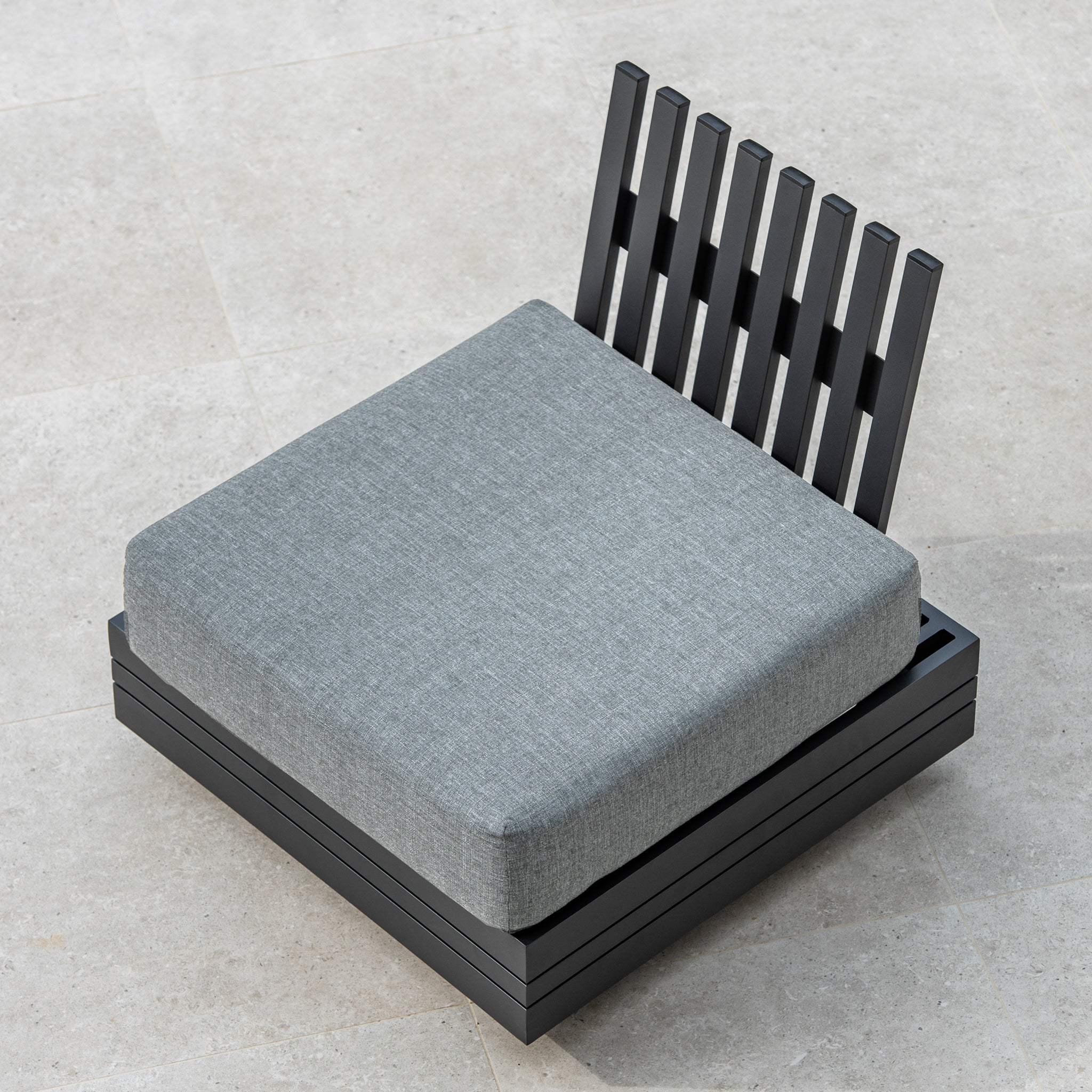 The Panama Corner Unit/Footstool in Washed Grey, featuring a stylish gray cushion and a sleek black slatted backrest, placed on a tiled surface, seamlessly integrates into any modular system.