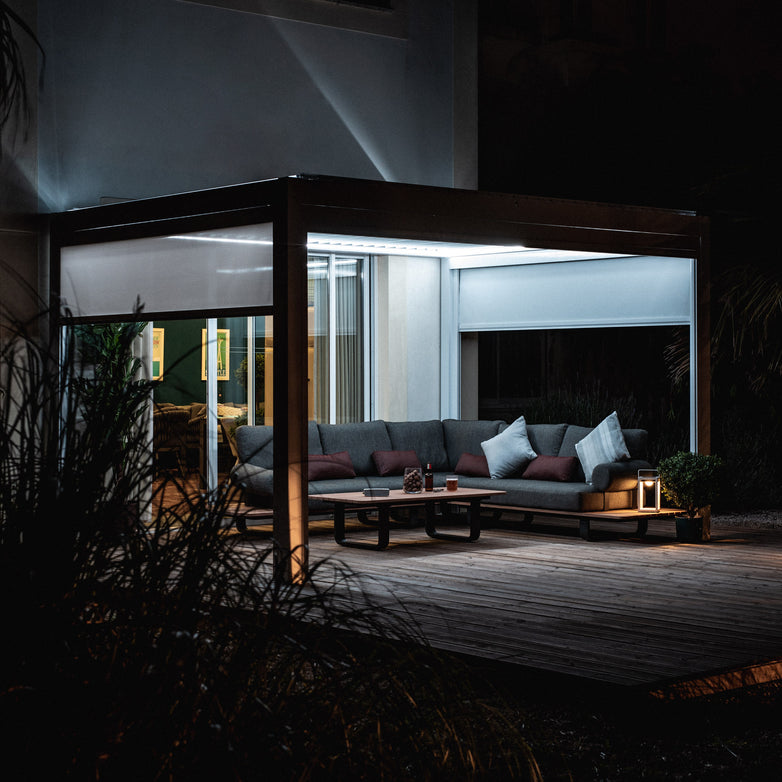 A modern, illuminated outdoor lounge at night featuring a sectional sofa, pillows, and a coffee table on a wooden deck with the PergoSTET 3m x 4m Rectangular Pergola in White, which includes 3 drop sides, LED lighting, and tilting louvres for adjustable shade.