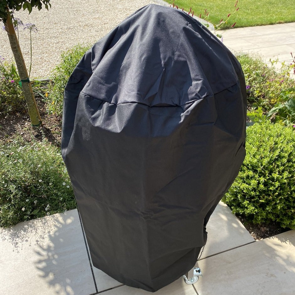 A black-covered Kamado grill stands in a garden on a patio, surrounded by greenery and a pebble path. The 18" Kamado grill, protected by its heat-resistant, waterproof Rain Cover for 18" Kamado Grills, blends seamlessly into its lush surroundings.