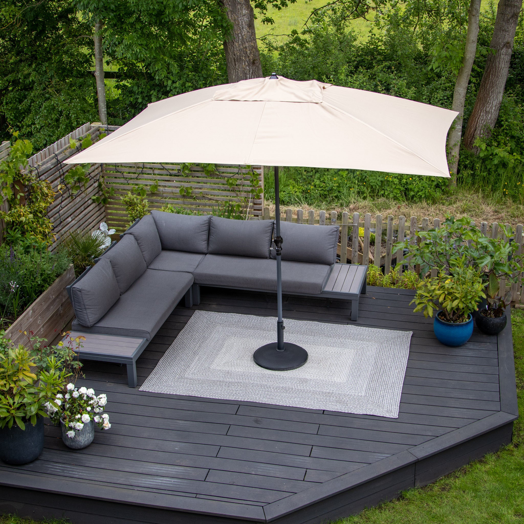 Aerial view of an outdoor patio featuring a weatherproof grey sectional sofa, beige umbrella, potted plants, and the Hestia 366cm x 274cm Weatherproof Indoor and Outdoor Rug in Walnut Grey on a wooden deck.