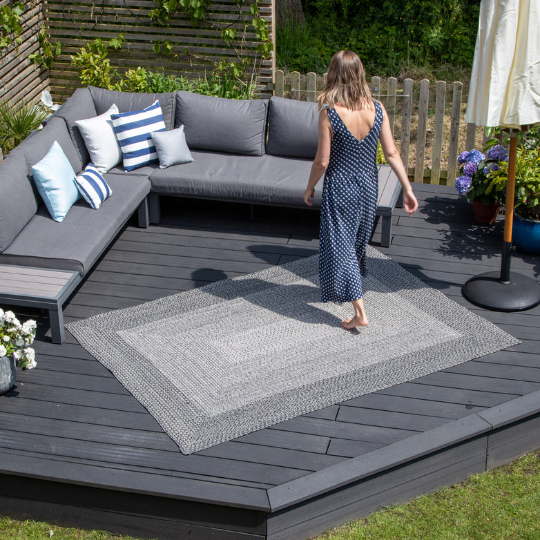 A woman in a polka dot dress walks barefoot on the Artemis 305cm x 244cm Weatherproof Indoor and Outdoor Rug in Slate Grey placed on a patio with outdoor seating and greenery around.