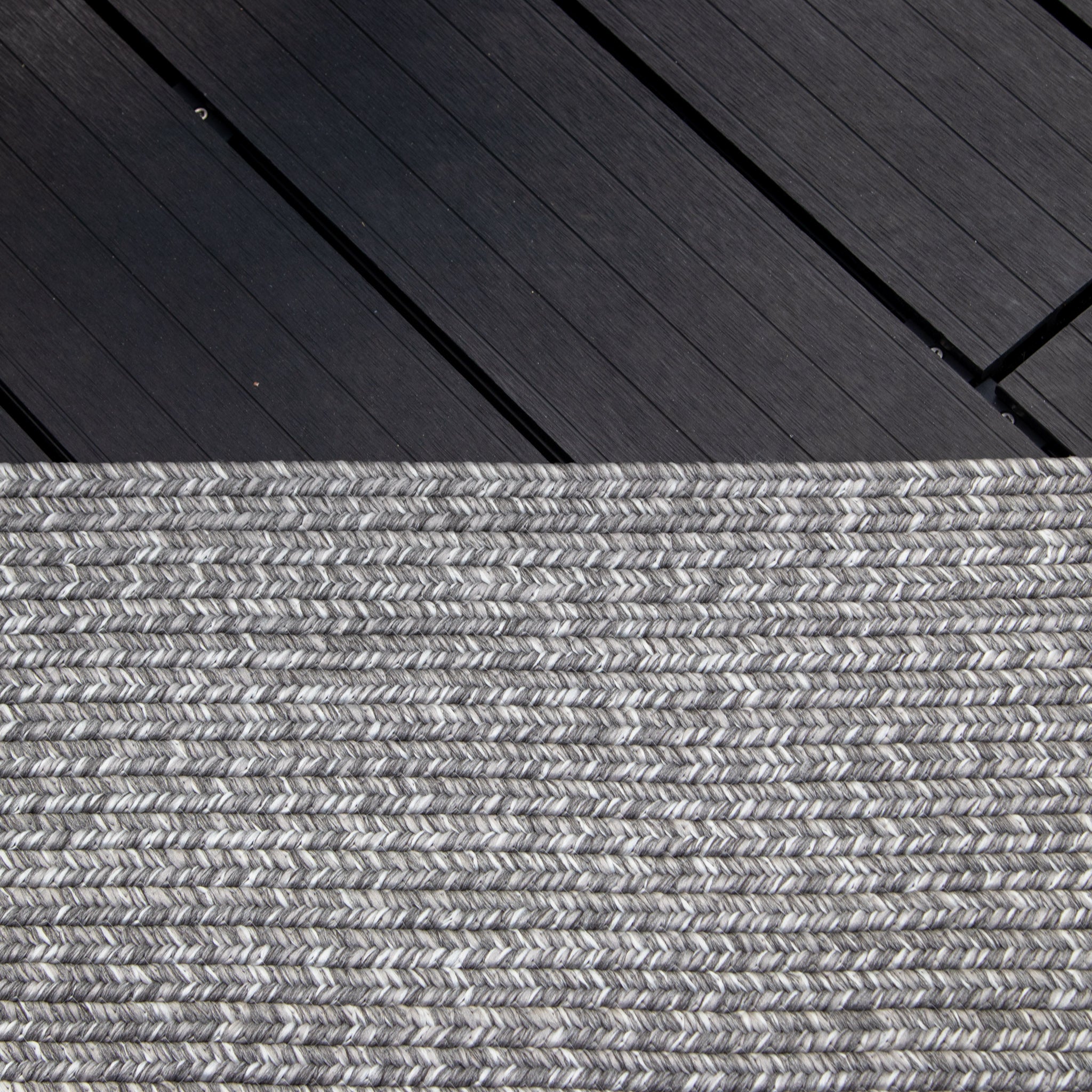 Close-up view of a dark wooden deck transitioning into the Hestia 366cm x 274cm Weatherproof Indoor and Outdoor Rug in Walnut Grey, UV resistant and weatherproof.