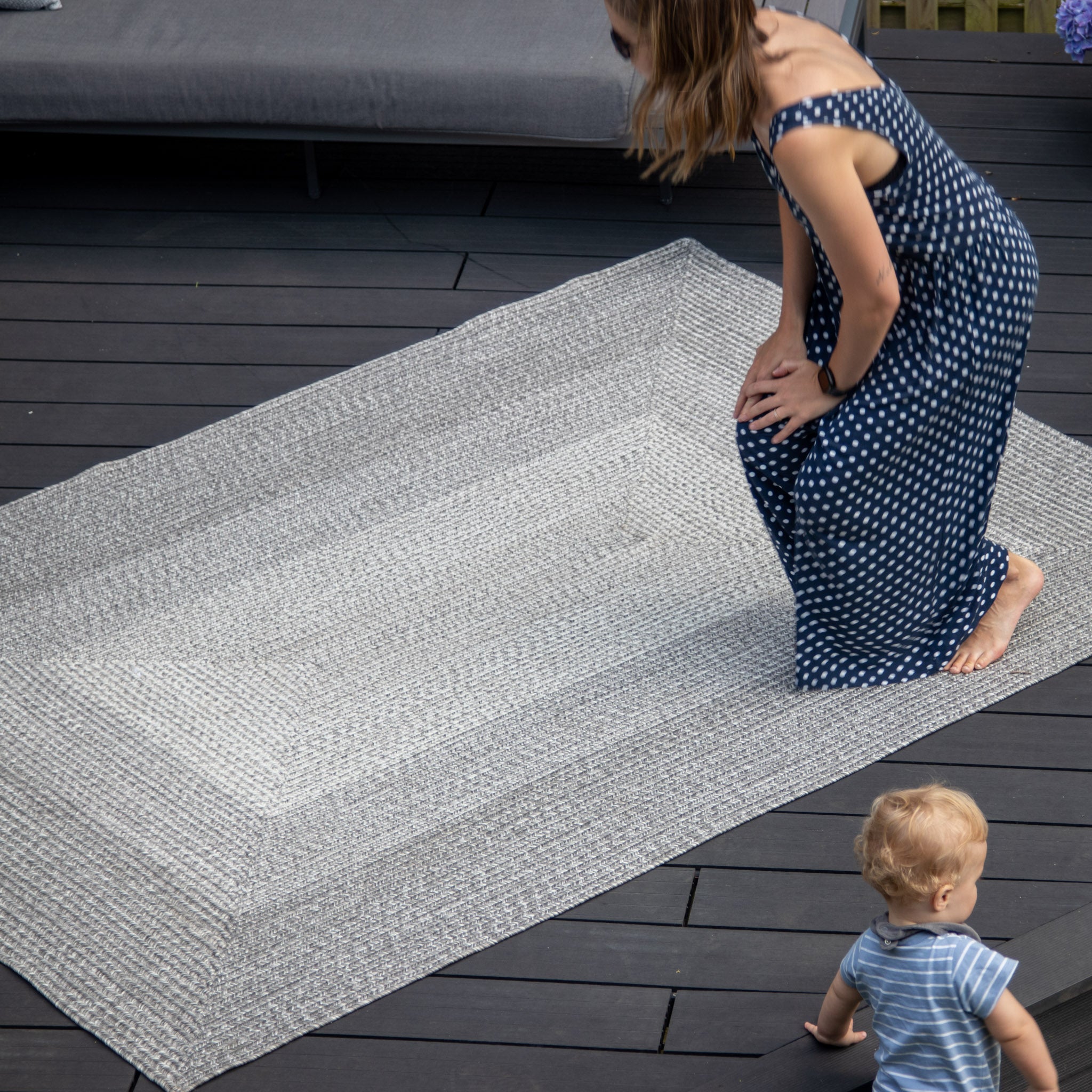 A woman in a blue dress adjusts a Hestia 366cm x 274cm Weatherproof Indoor and Outdoor Rug in Walnut Grey on a wooden deck, while a small child in a striped outfit watches.