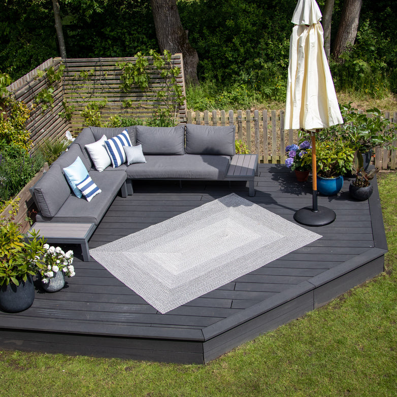 A cozy outdoor patio features Walnut Grey seating, blue-striped pillows, a spacious Hestia 290cm x 190cm Weatherproof Indoor and Outdoor Rug in Walnut Grey, potted plants, and an umbrella in a green garden. The UV-resistant components ensure longevity and comfort throughout the seasons.