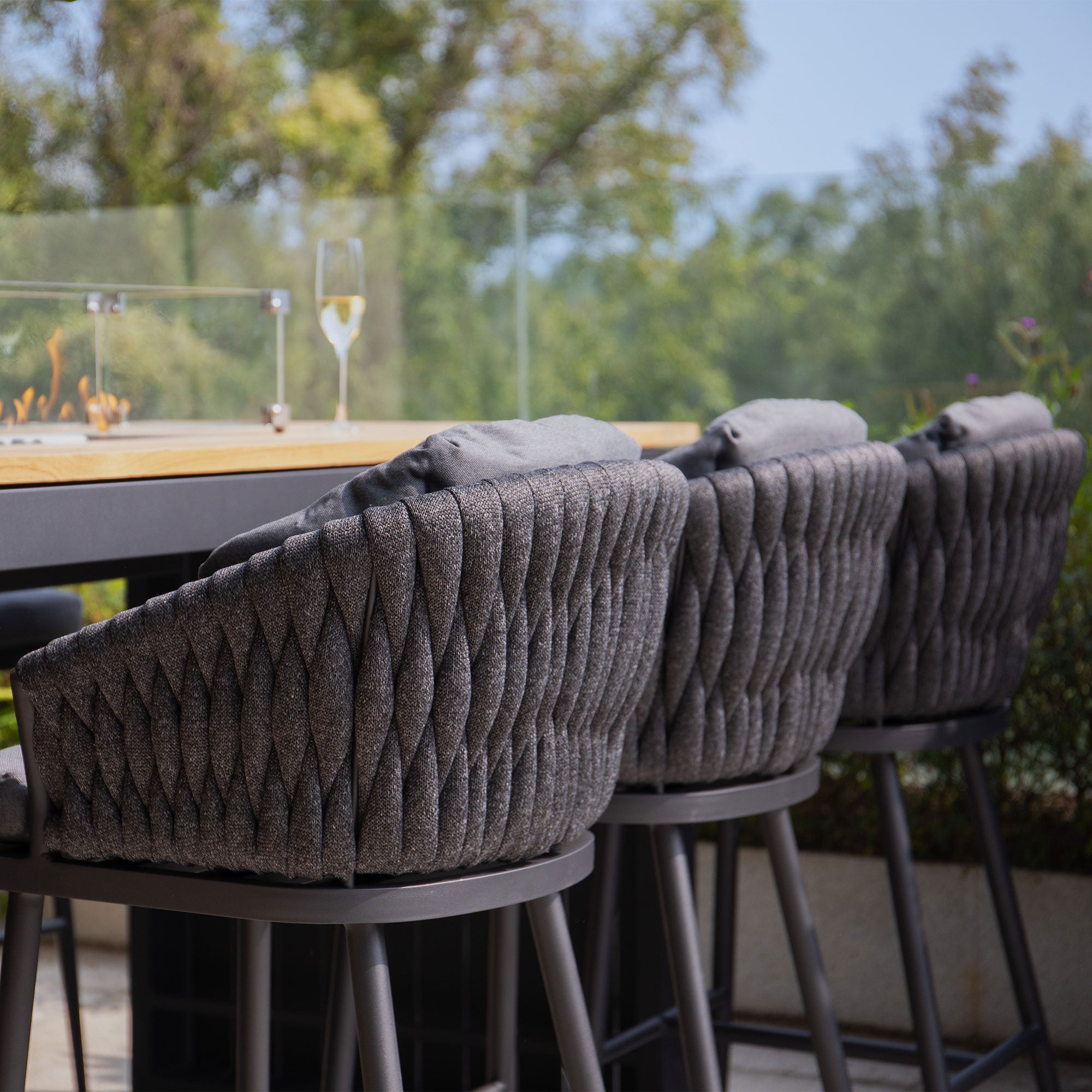 Three cushioned bar stools from the Palma 6 Seat Rope Teak Firepit Bar Set in Grey are lined up at a high outdoor table with a glass of white wine, set in a garden setting.